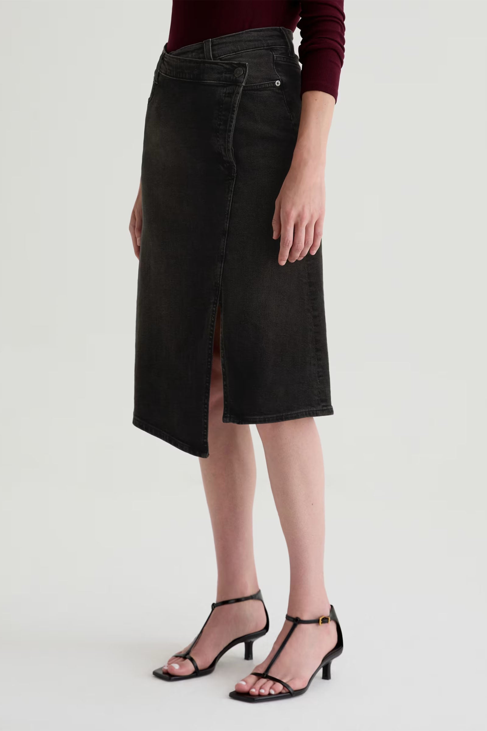 A person wears the Bruna Wrap Midi Skirt—dark denim, knee-length with an asymmetric front slit—paired with a burgundy long-sleeve top and black open-toe heeled sandals, standing against a plain background.