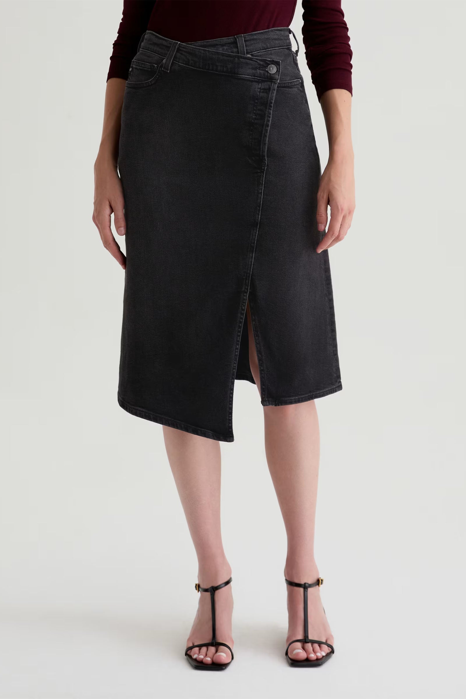 A person wearing the Bruna Wrap Midi Skirt in dark gray with an asymmetrical design and front slit, styled with black strappy sandals, stands against a plain light background.