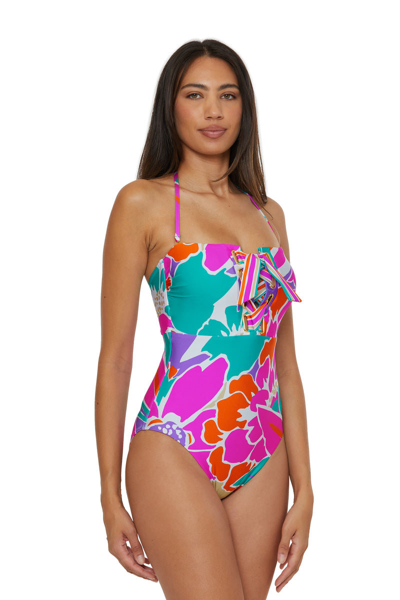A woman models the Lace-Up Bandeau One-Piece Swimsuit - Amalfi, featuring a bold floral print in pink, orange, teal, and green with a front tie detail and halter neck strap, against a plain white background.