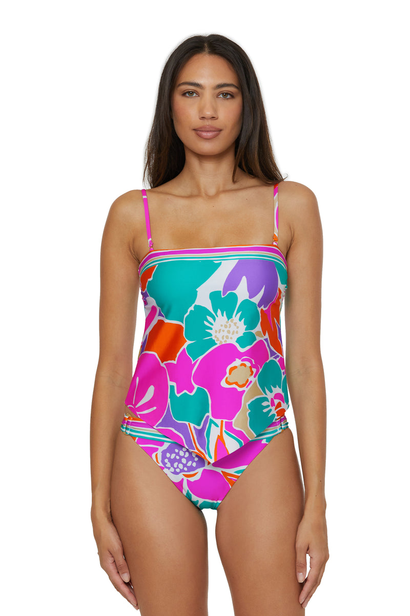 A woman with long dark hair models the TRINA TURK SWIM Handkerchief Tankini Swim Top - Amalfi, featuring bold, vibrant floral patterns in pink, teal, purple, and orange. She stands facing forward against a white background.