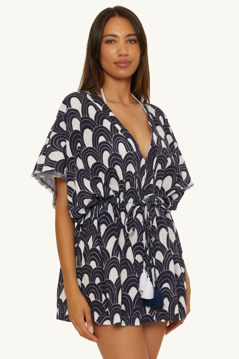A woman wears the V-Neck Cover-up Tunic Dress - Arches, featuring a short, loose fit with a deep V-neck, navy and white arches pattern, drawstring waist, and tassel ties, posed against a plain light background.