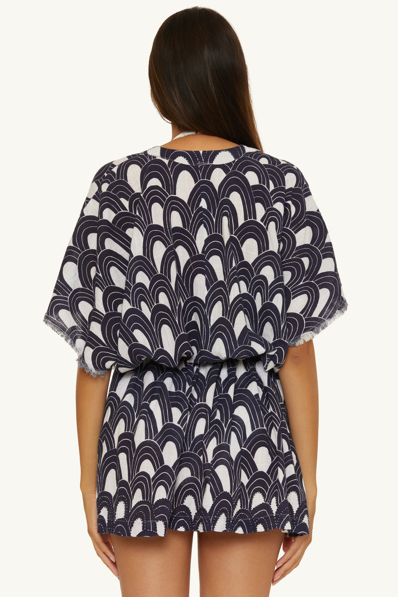 A woman with long brown hair faces away, modeling the V-Neck Cover-up Tunic Dress - Arches, a short, loose-fitting style with wide sleeves and a bold navy and white scallop pattern. The background is plain white.