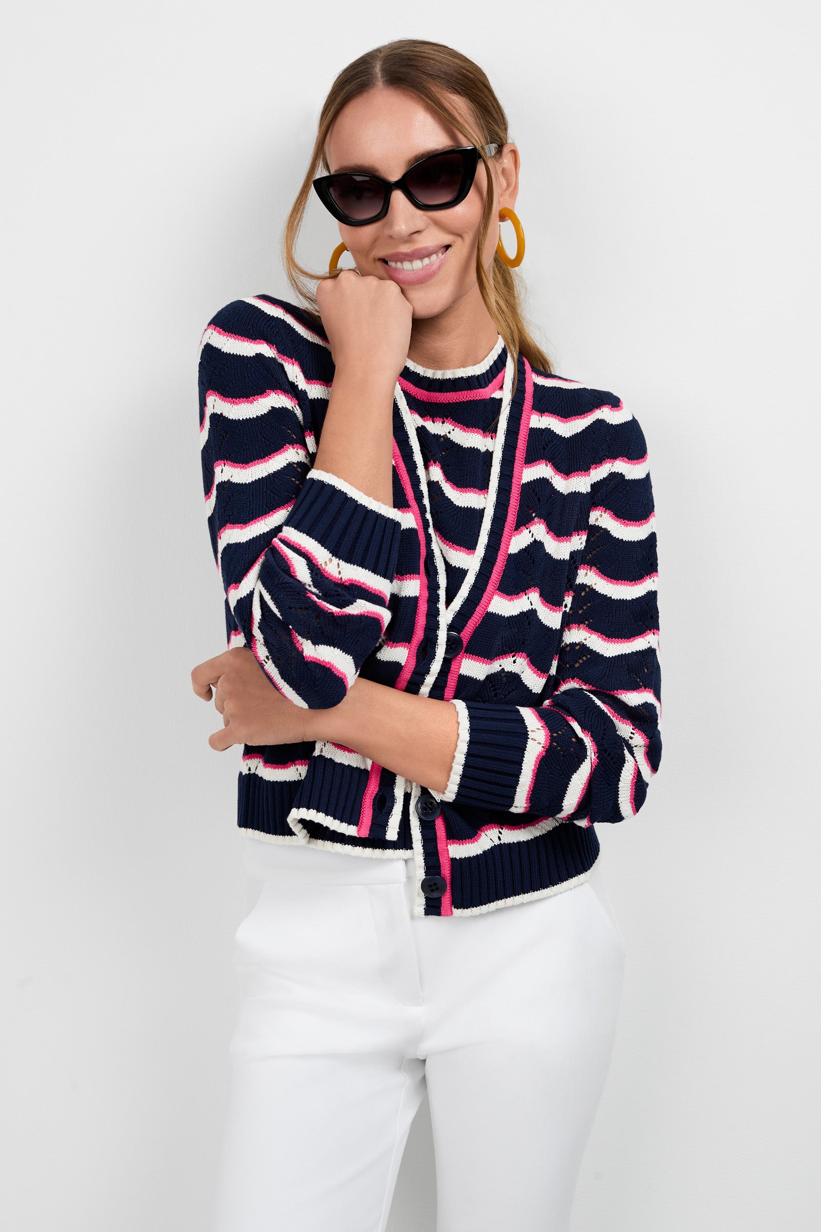 A woman smiles against a white background, wearing cat-eye sunglasses, gold hoop earrings, white pants, and the Alessia Knit Cardigan—a navy blue, pink, and white striped sweater—with one arm crossed and the other hand on her chin.