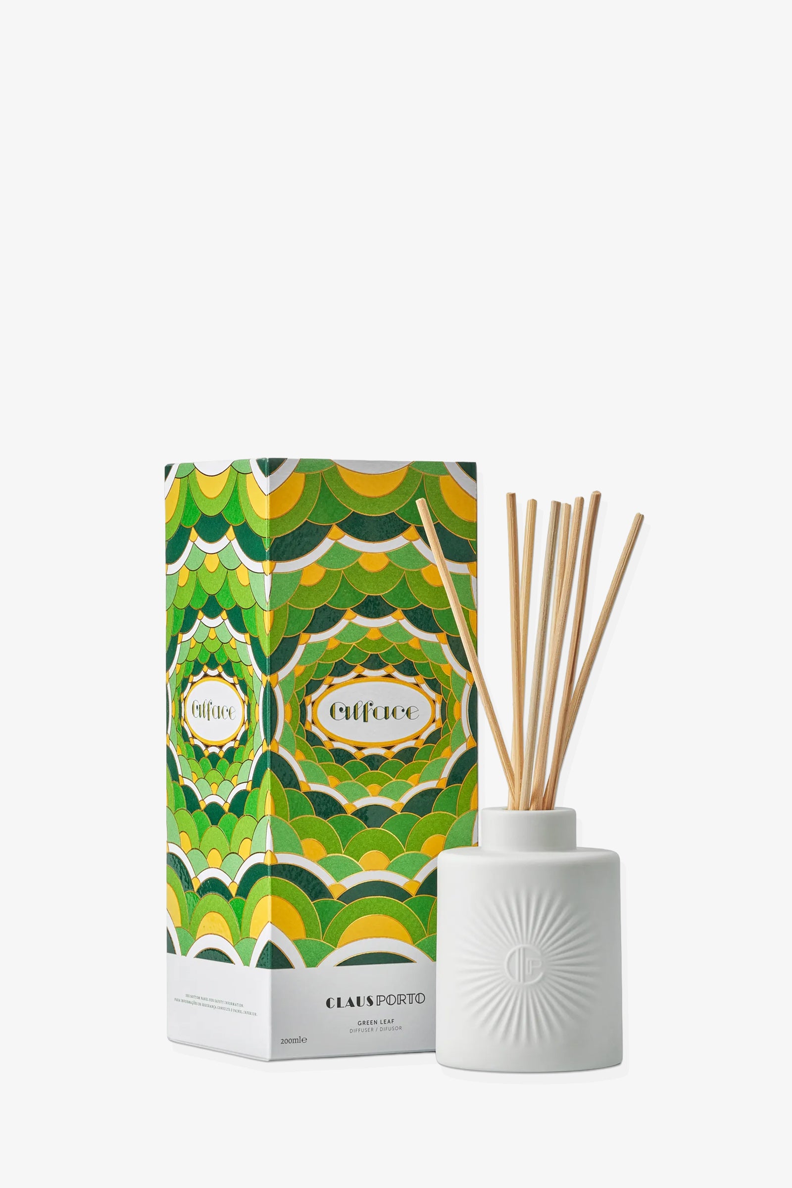 A white ceramic diffuser with reed sticks stands next to a vibrant green, yellow, and white patterned Claus Porto Alface Diffuser box.