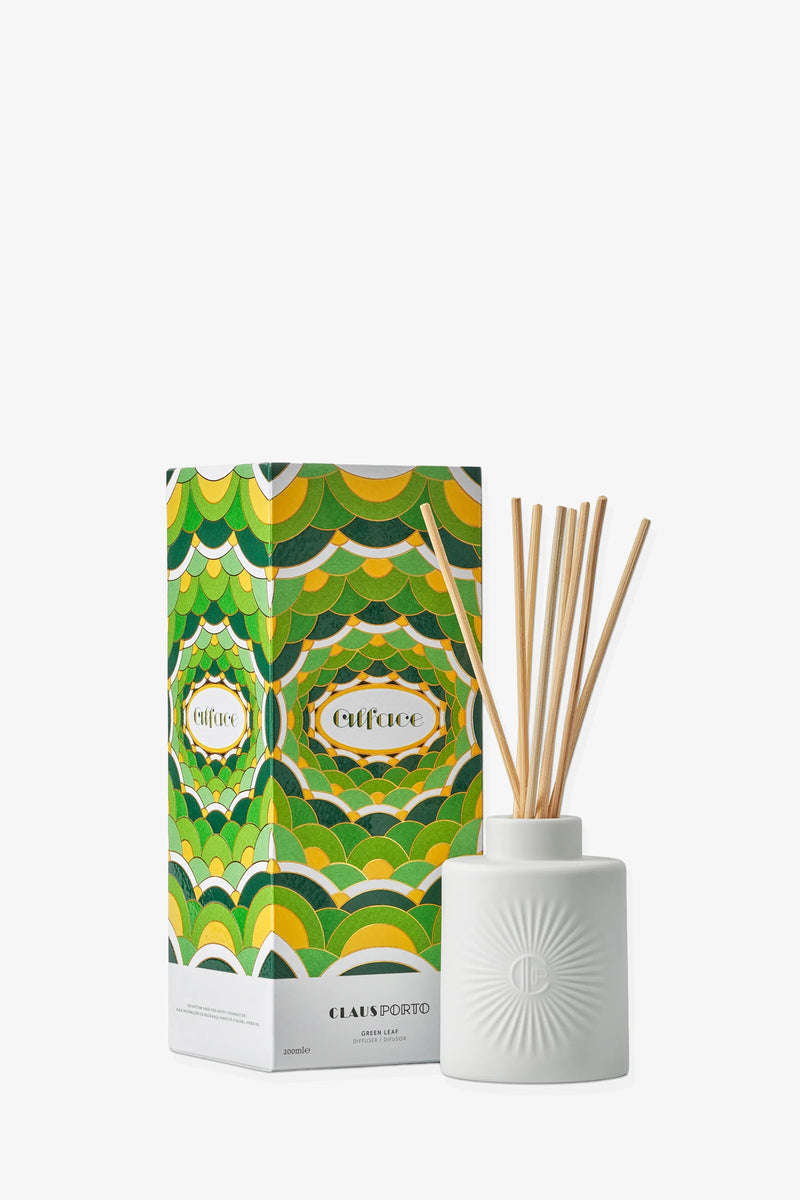 A white ceramic diffuser with reed sticks stands next to a vibrant green, yellow, and white patterned Claus Porto Alface Diffuser box.