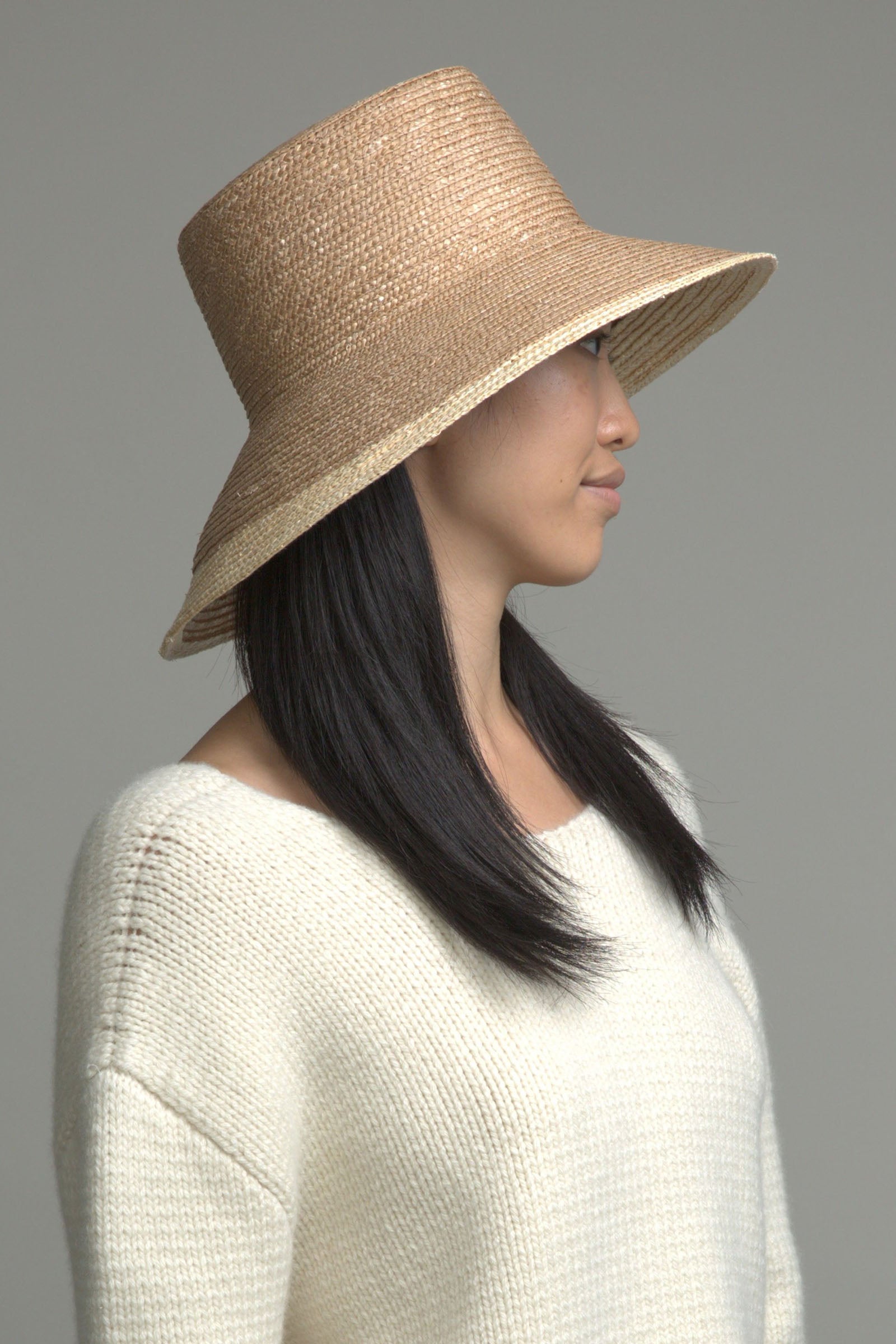 A woman with long, straight black hair wears the Eugenia Kim Annabelle Hat and a cream knit sweater, facing sideways against a plain gray background.