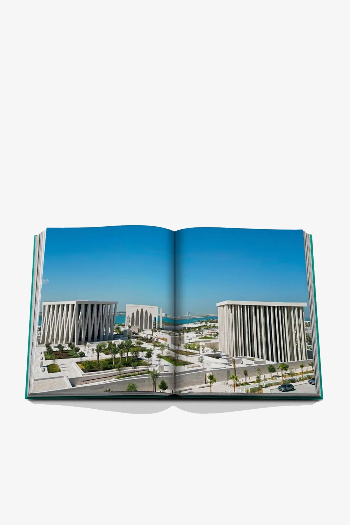 An open Assouline Abu Dhabi Book displays a photograph of modern, white rectangular buildings with columns set against a blue sky and water, capturing an urban architectural landscape.