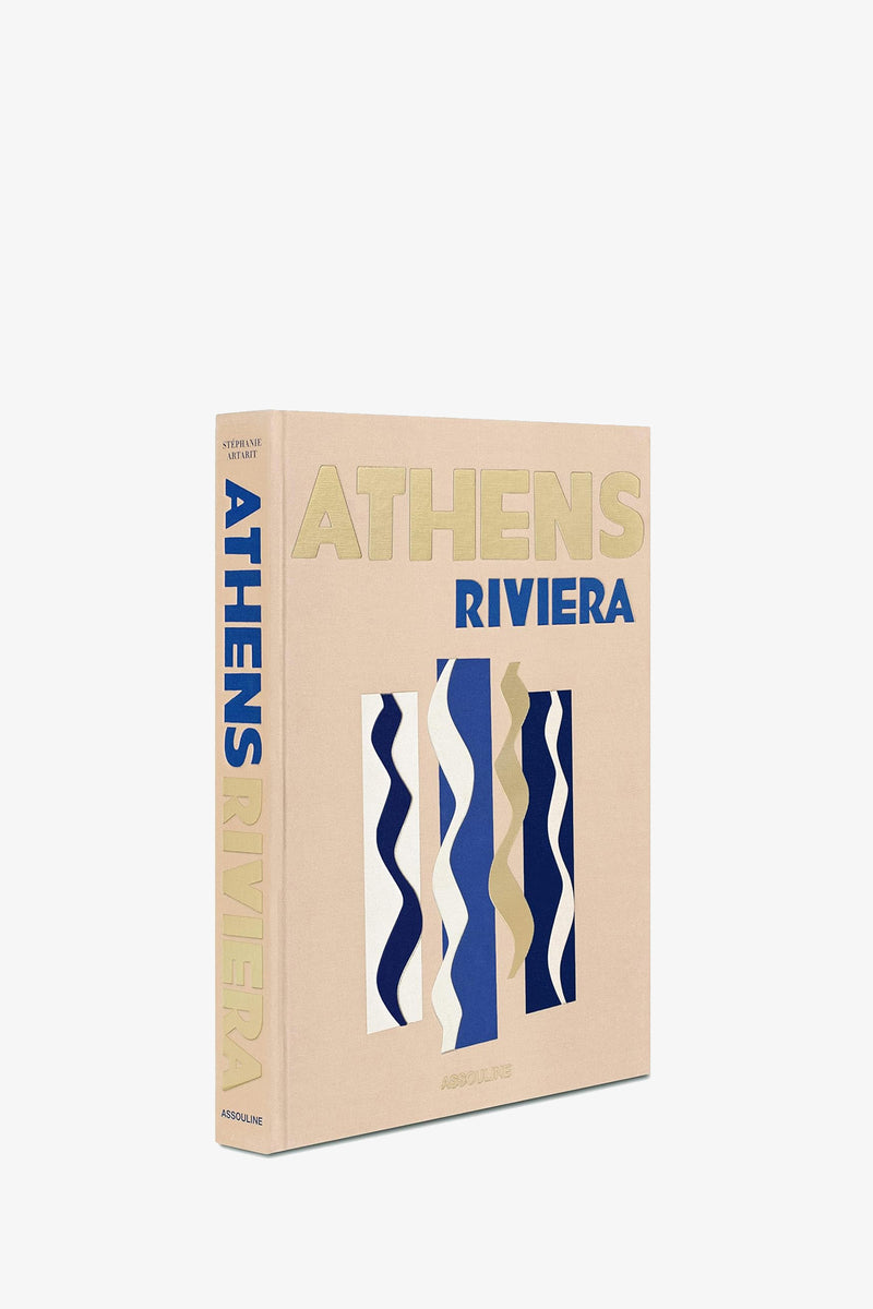 An Assouline Athens Riviera Book, in beige with gold and blue lettering, stands upright against a white background. Its cover features abstract wavy shapes in blue, white, and beige.
