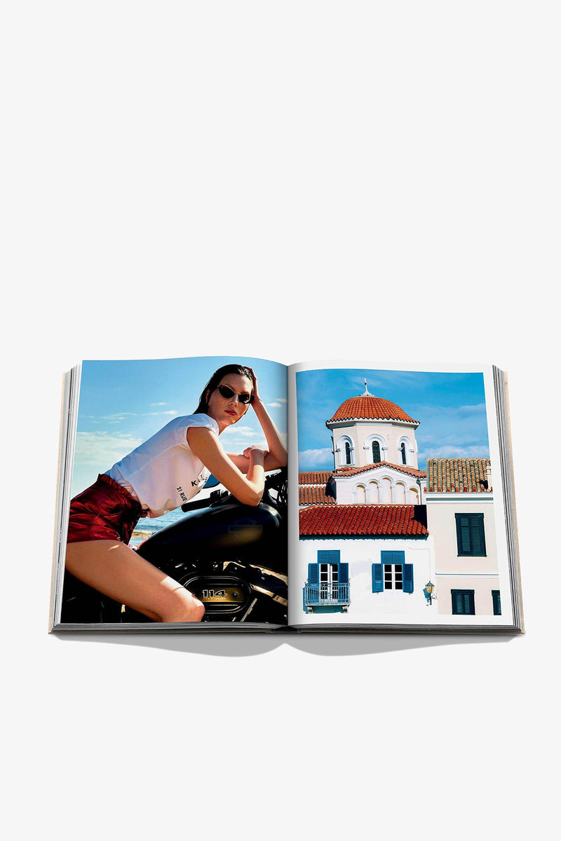 An open copy of the Assouline Athens Riviera Book displays a photo of a woman in sunglasses and shorts on the left page, and an image of a white building with a red-tiled dome set against a blue sky on the right page.