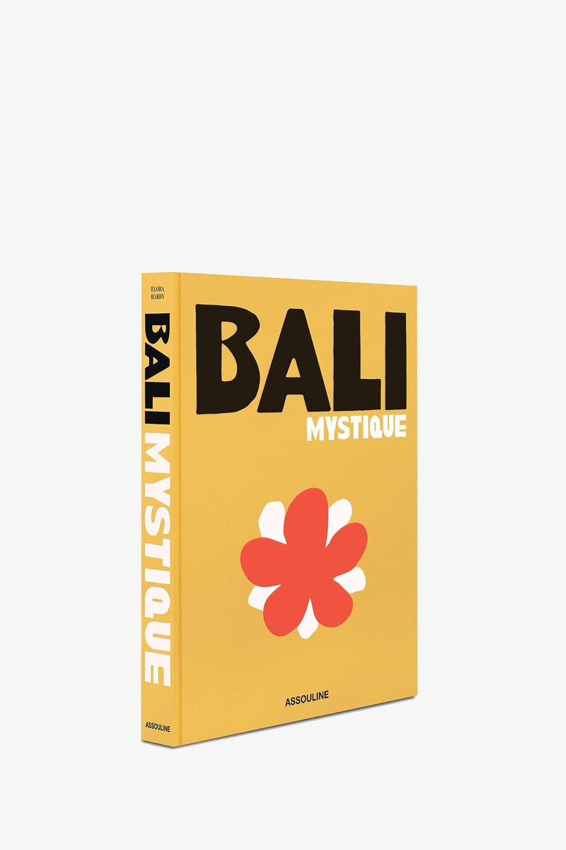 A yellow book titled ASSOULINE BALI MYSTIQUE featuring a large red and white flower on the cover, with the title also appearing on the spine and Assouline printed at the bottom.