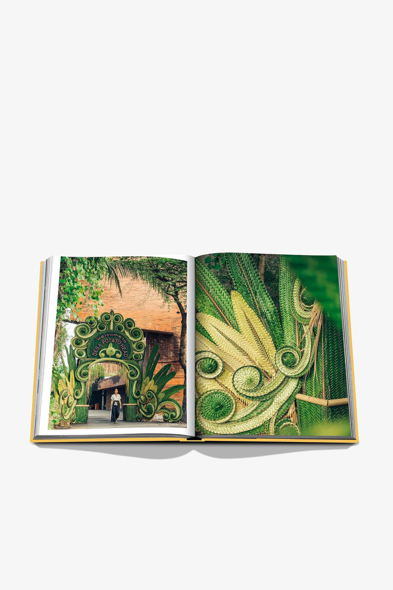 An open ASSOULINE BALI MYSTIQUE book displays a colorful outdoor archway and intricate green leaf designs, with two people walking under the archway on the left page.