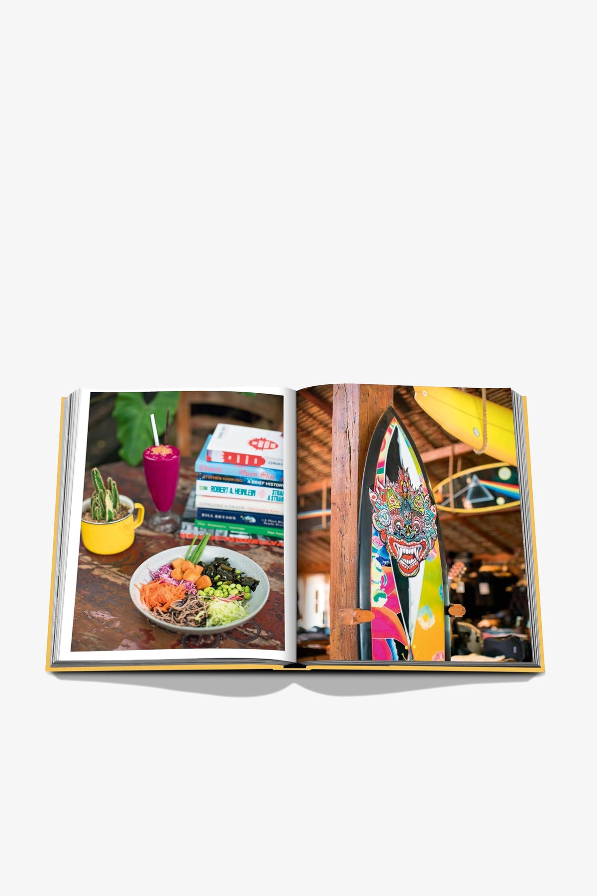 An open ASSOULINE BALI MYSTIQUE book displays a vibrant photo of a colorful meal with drinks and stacked books on one page, while the opposite page features an image of a decorated surfboard inside a rustic, lively cafe.