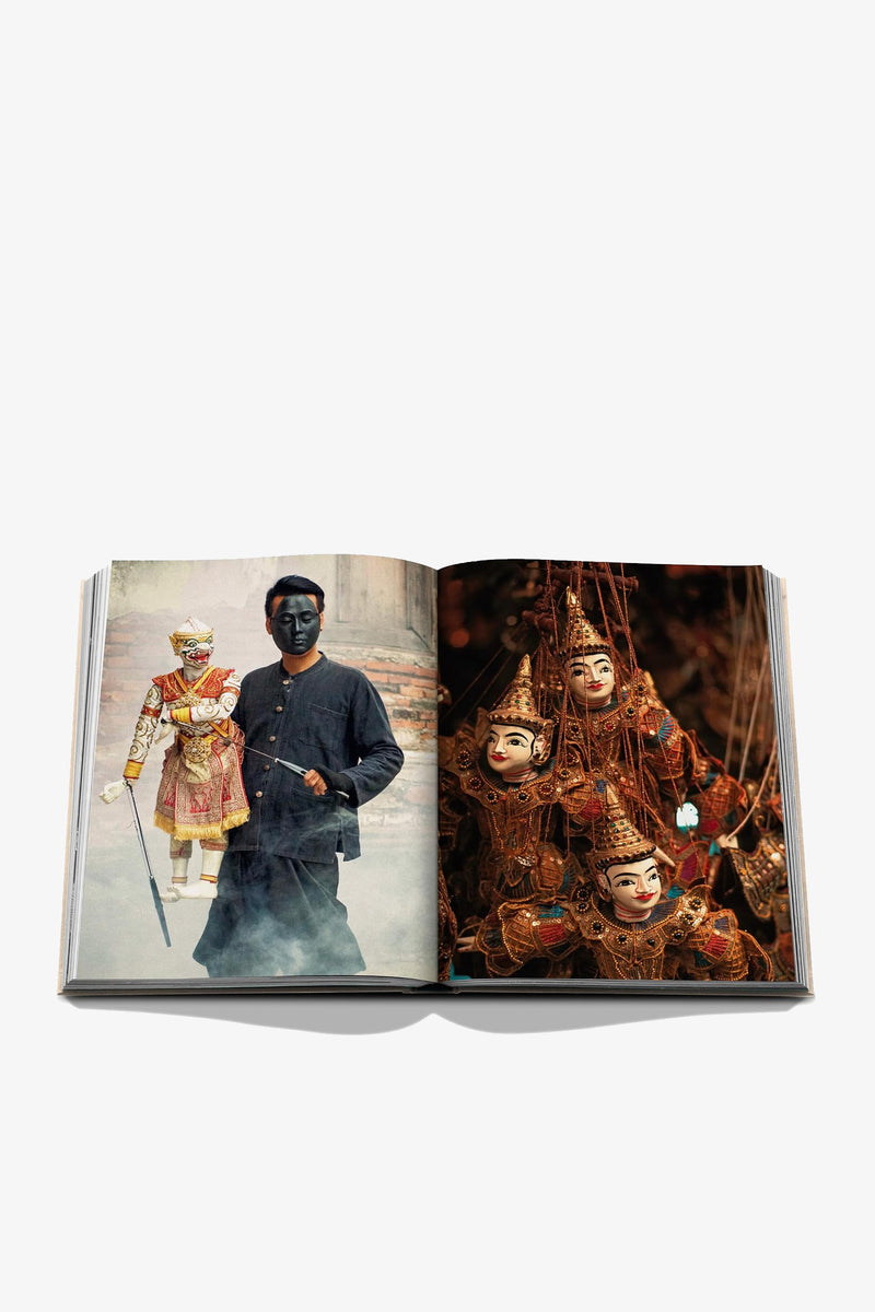 The Assouline Bangkok Spirit Book is open to a spread showing a masked figure holding a puppet on the left page, while the right page features an array of ornate puppets with expressive faces and elaborate costumes, all set against a white background.