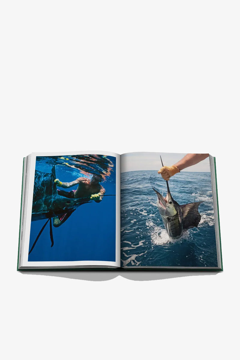 An open Assouline Costa Rica Vida Book displays two ocean photographs: on the left, a person swims underwater; on the right, a gloved hand lifts a large, long-billed fish above the water.