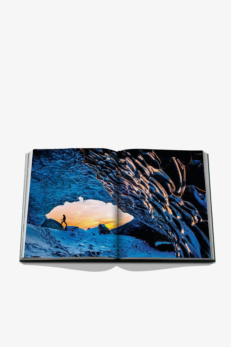An open Assouline Iceland Epic Book displays a vivid photo of a person walking in a blue ice cave with a sunset sky visible through the cave’s opening.