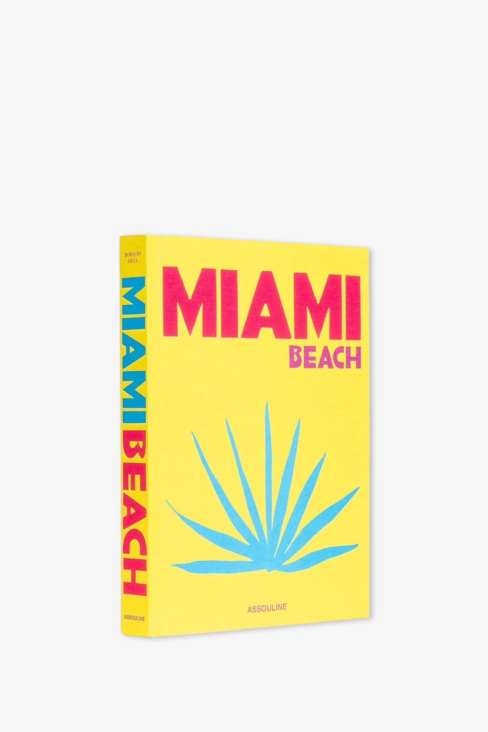 A yellow Assouline Miami Beach Book featuring the title “MIAMI BEACH” in bold pink and blue letters with a blue palm graphic on the cover; the spine displays “MIAMI BEACH” vertically in matching pink and blue.