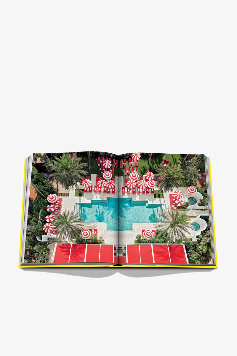 An open Assouline Miami Beach Book displays an aerial photo of a pool surrounded by palm trees, red-and-white umbrellas, sun loungers, and a vibrant outdoor setting.
