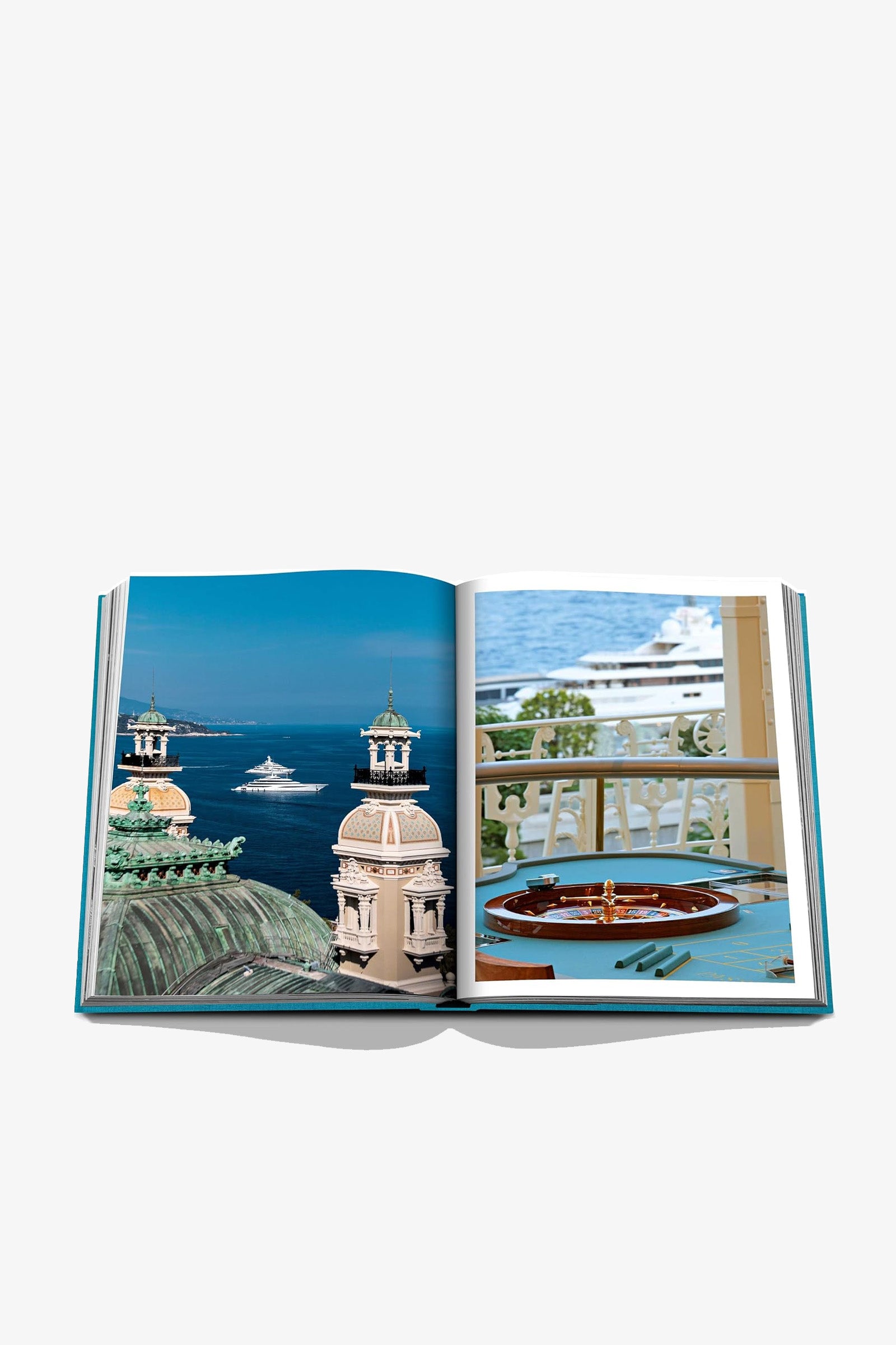 An open Assouline Monte Carlo Book displays a photo of a picturesque coastline with elegant towers and yachts, alongside another image featuring a balcony overlooking the sea with a table set with plates and cutlery.