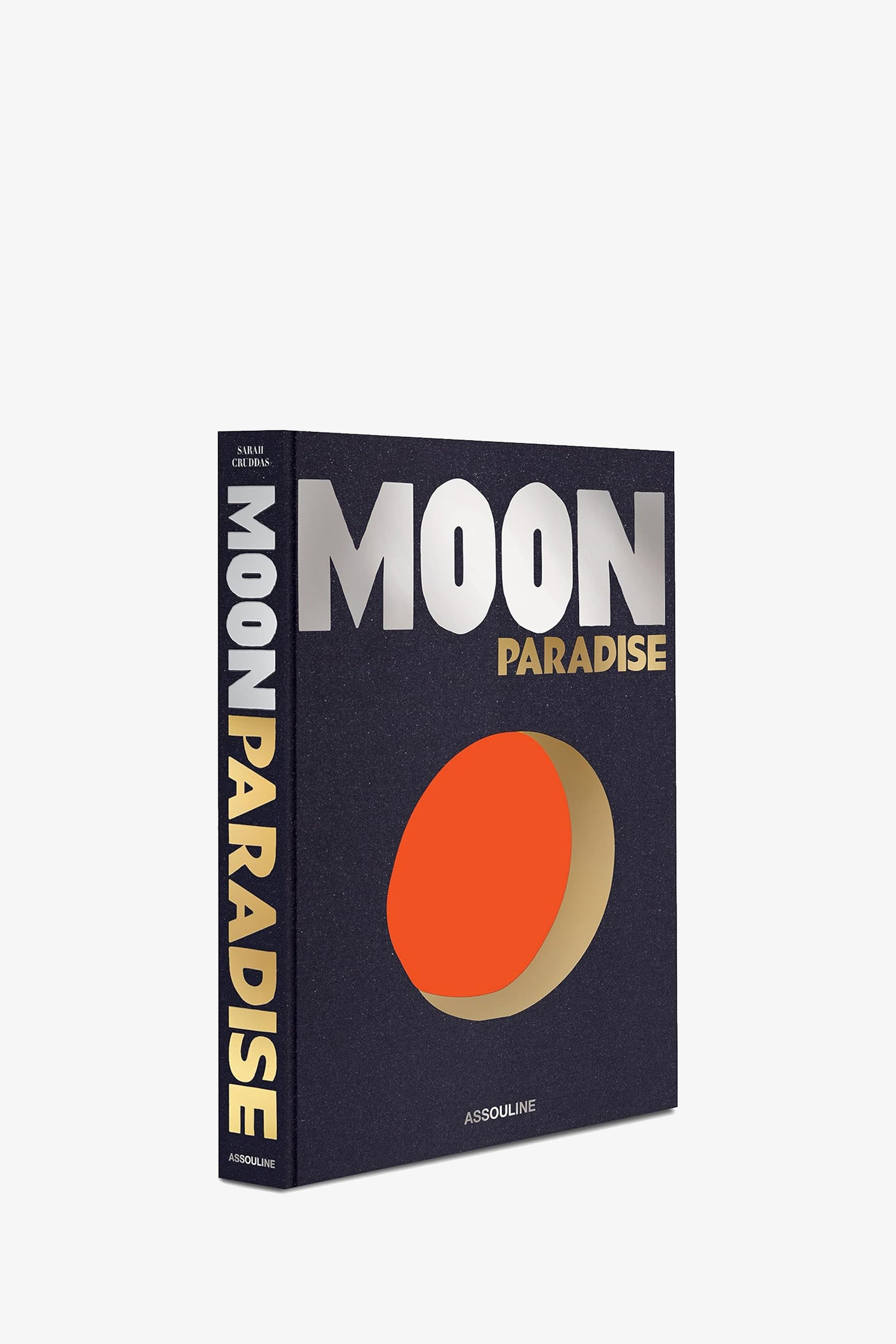 The Assouline Moon Paradise Book features metallic silver and gold text on a dark cover, with a stylized orange and beige moon graphic, and the title displayed in large vertical letters along the spine.