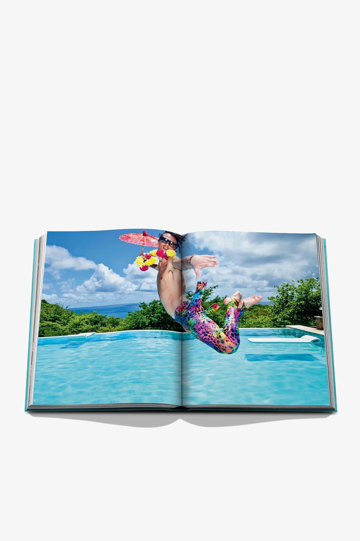 An open copy of the Assouline Mustique Icon Book displays a photo featuring a person in colorful swimwear jumping into a pool, holding a drink and an inflatable toy, with lush greenery and blue sky in the background.