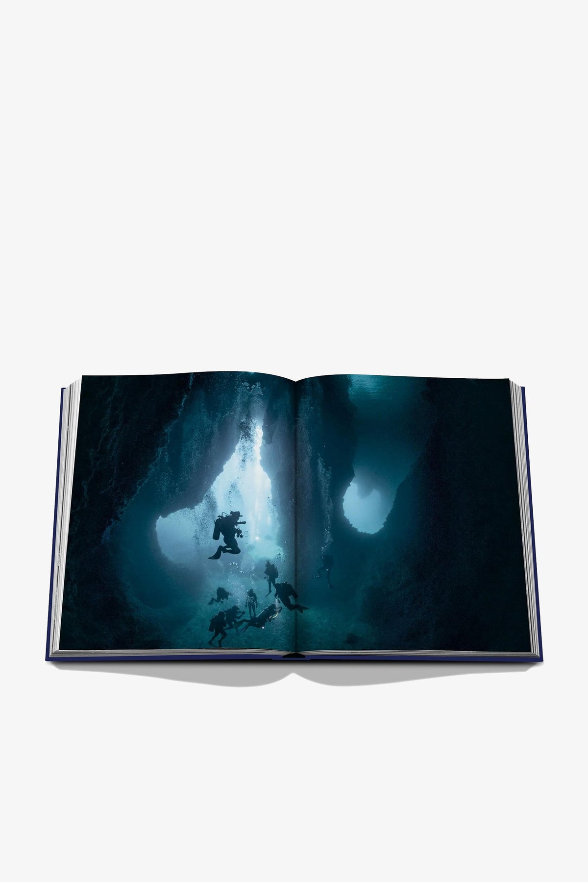 An open copy of Assouline Ocean Wanderlust displaying a photograph of scuba divers exploring an underwater cave, with beams of light filtering through the water.