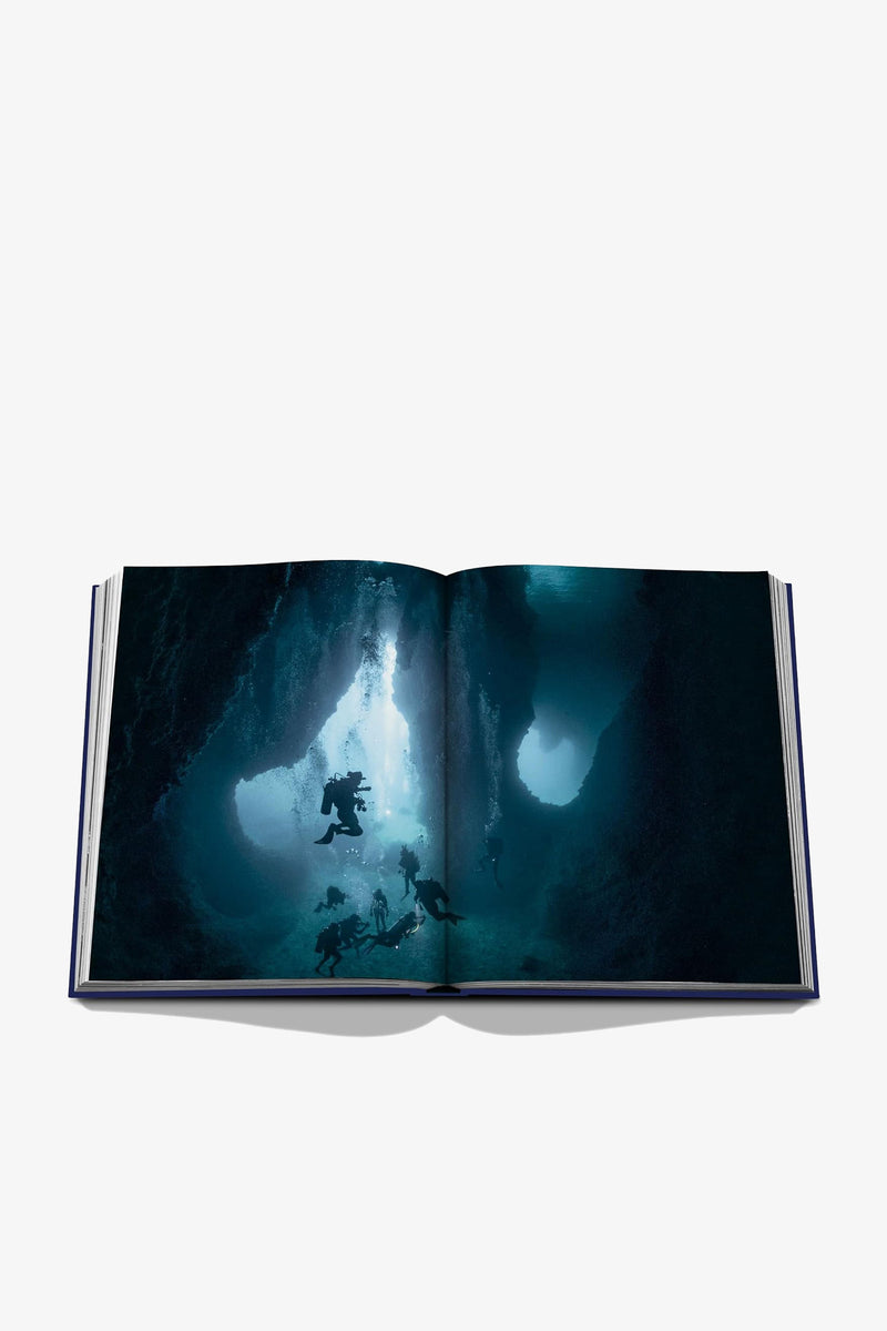 An open copy of Assouline Ocean Wanderlust displaying a photograph of scuba divers exploring an underwater cave, with beams of light filtering through the water.