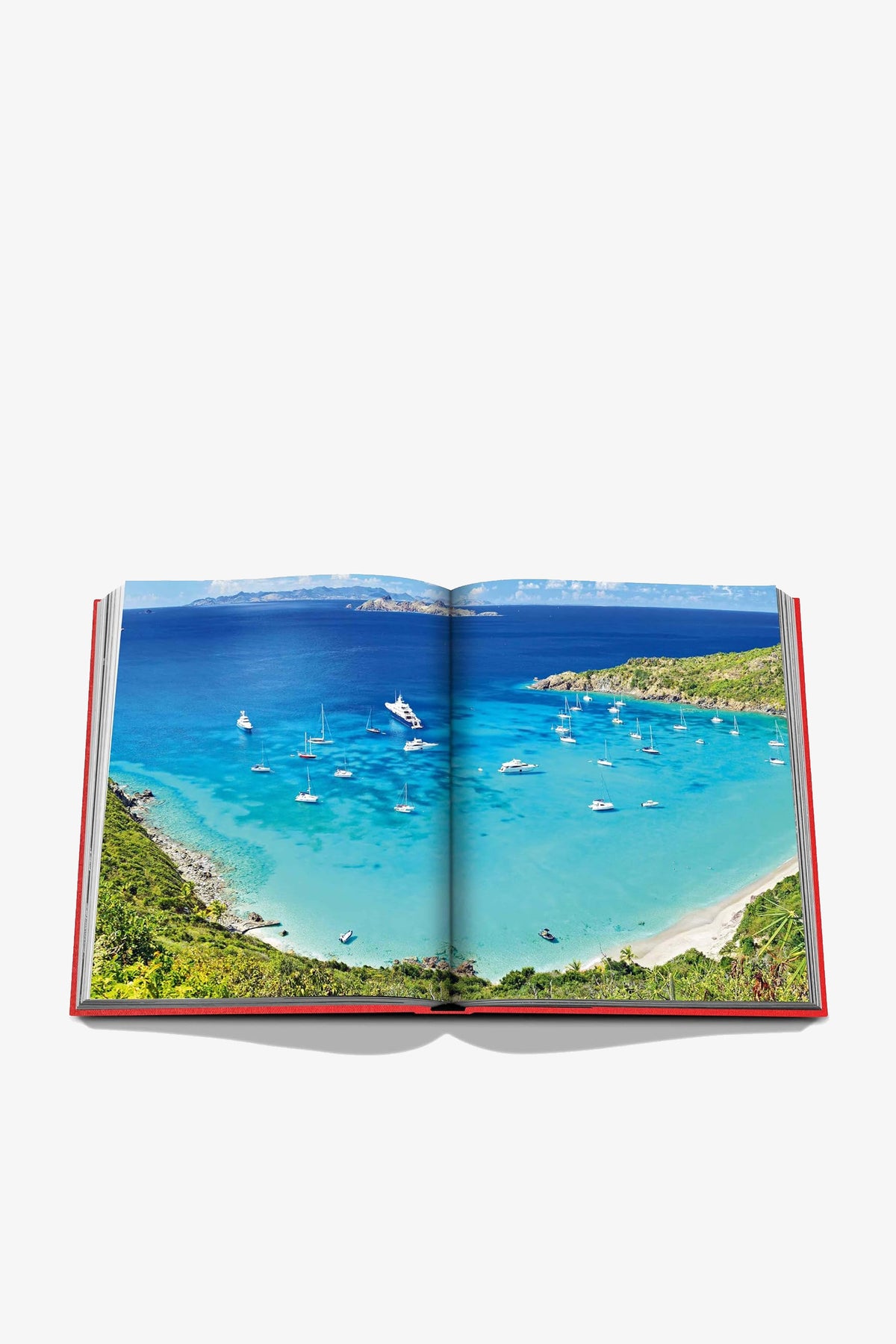 An open Assouline St Barths Freedom Book displays a vivid photograph of a tropical bay with turquoise water, sailboats, green hills, and a clear blue sky.