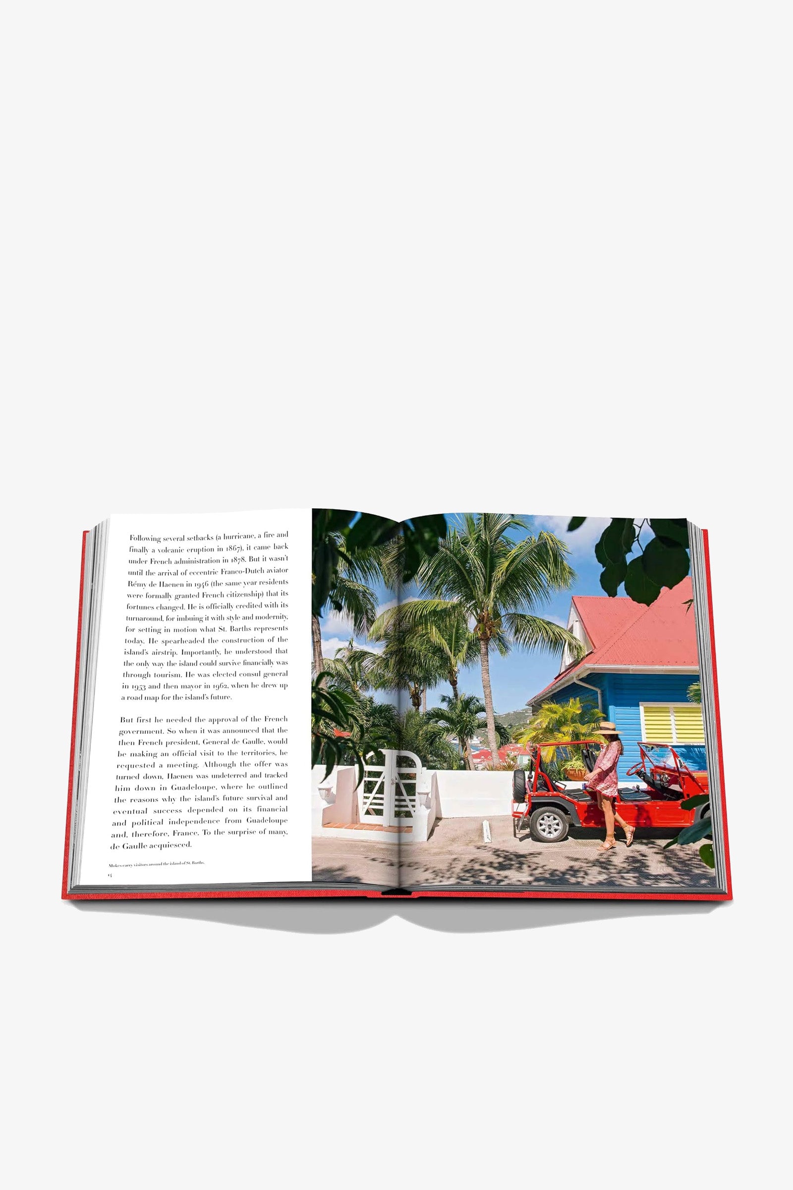 The Assouline St Barths Freedom Book is open on a white background, displaying a page of text on the left and, on the right, a vibrant photo featuring palm trees, a yellow cottage, and a red convertible with two people.