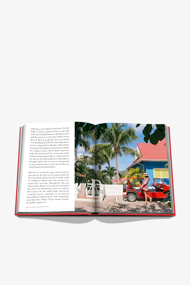 The Assouline St Barths Freedom Book is open on a white background, displaying a page of text on the left and, on the right, a vibrant photo featuring palm trees, a yellow cottage, and a red convertible with two people.