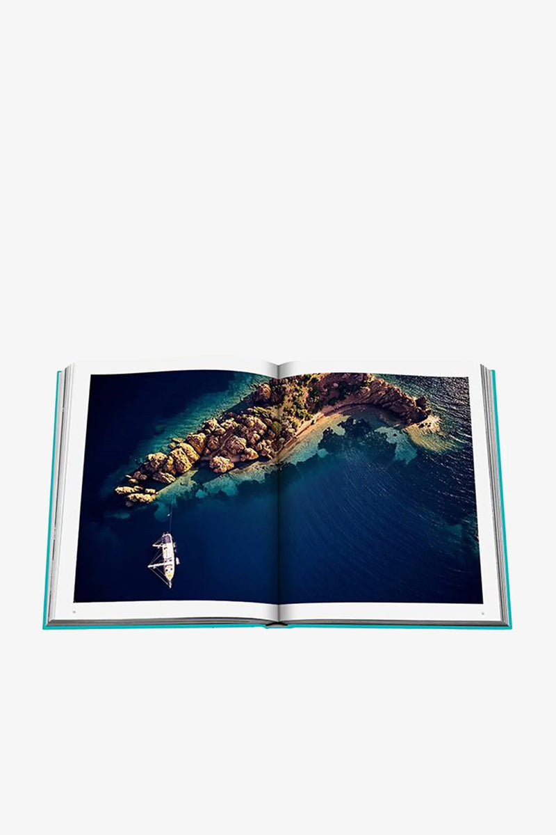An open Assouline Turquoise Coast Book displays an aerial photo of a rocky island surrounded by blue water, with a sailboat nearby.