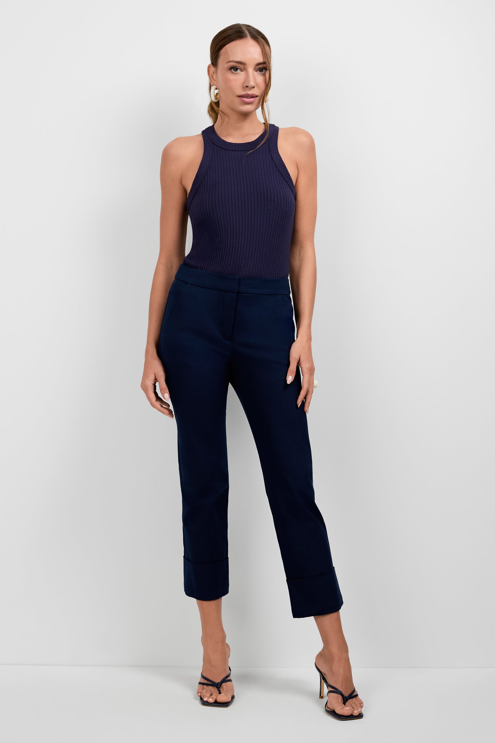 A woman stands against a plain light wall, wearing sleeveless navy ribbed top, Banshee Cropped Cuff Hem Pants, and black heeled sandals. Her light brown hair is pulled back and she accessorizes with hoop earrings.
