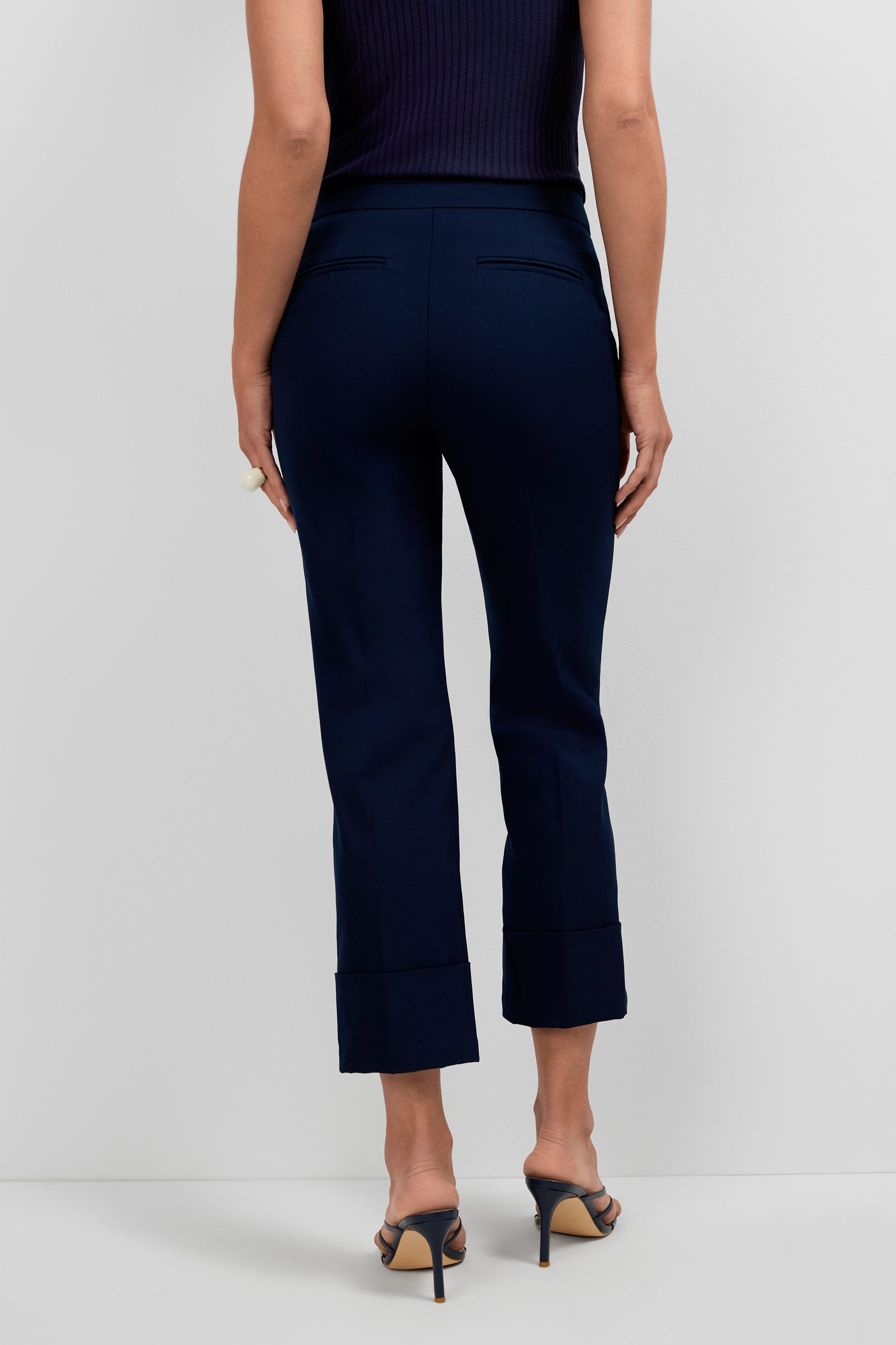 A person shown from the back wears Banshee Cropped Cuff Hem Pants in navy blue with cuffed hems and welt pockets, styled with heeled sandals and a sleeveless black top.
