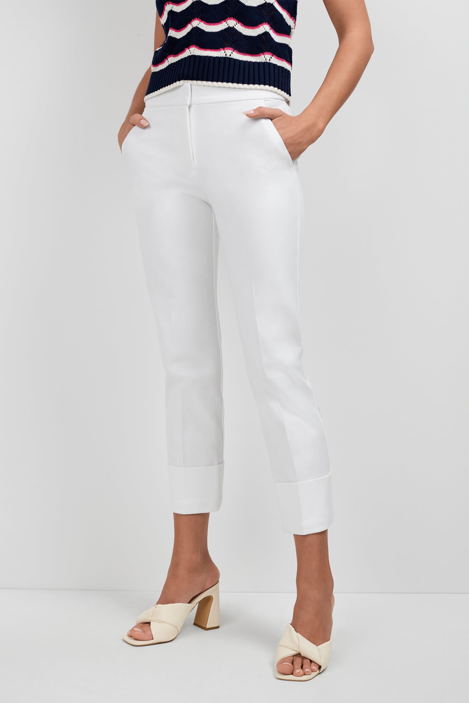 A person stands against a white wall, wearing Banshee Cropped Cuff Hem Pants, a sleeveless patterned top, and cream heeled sandals with crossed straps, hands in their pockets.