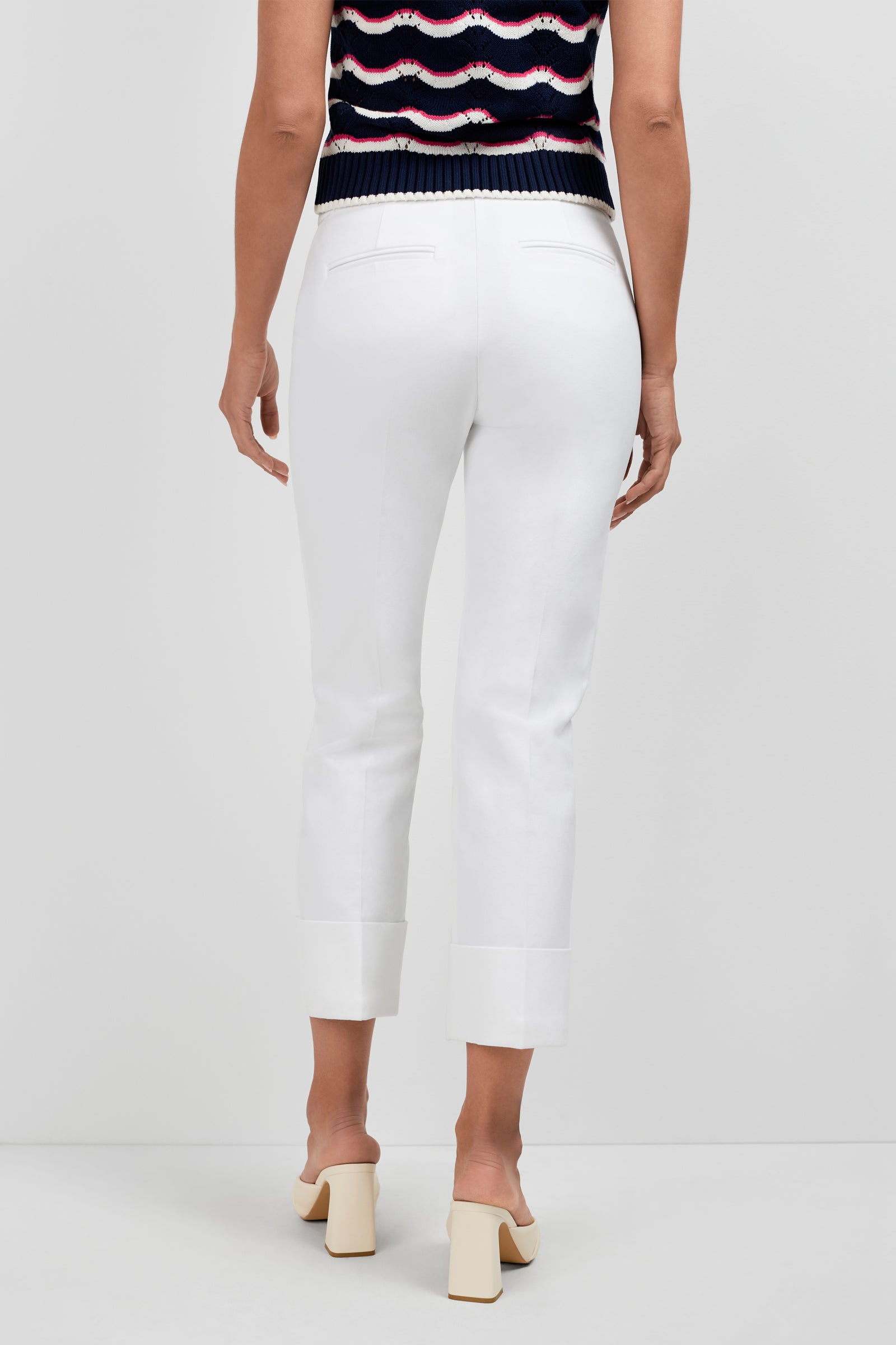 A person stands facing away against a plain white background, wearing Banshee Cropped Cuff Hem Pants, a sleeveless navy top with red and white stripes, and cream heeled mules.