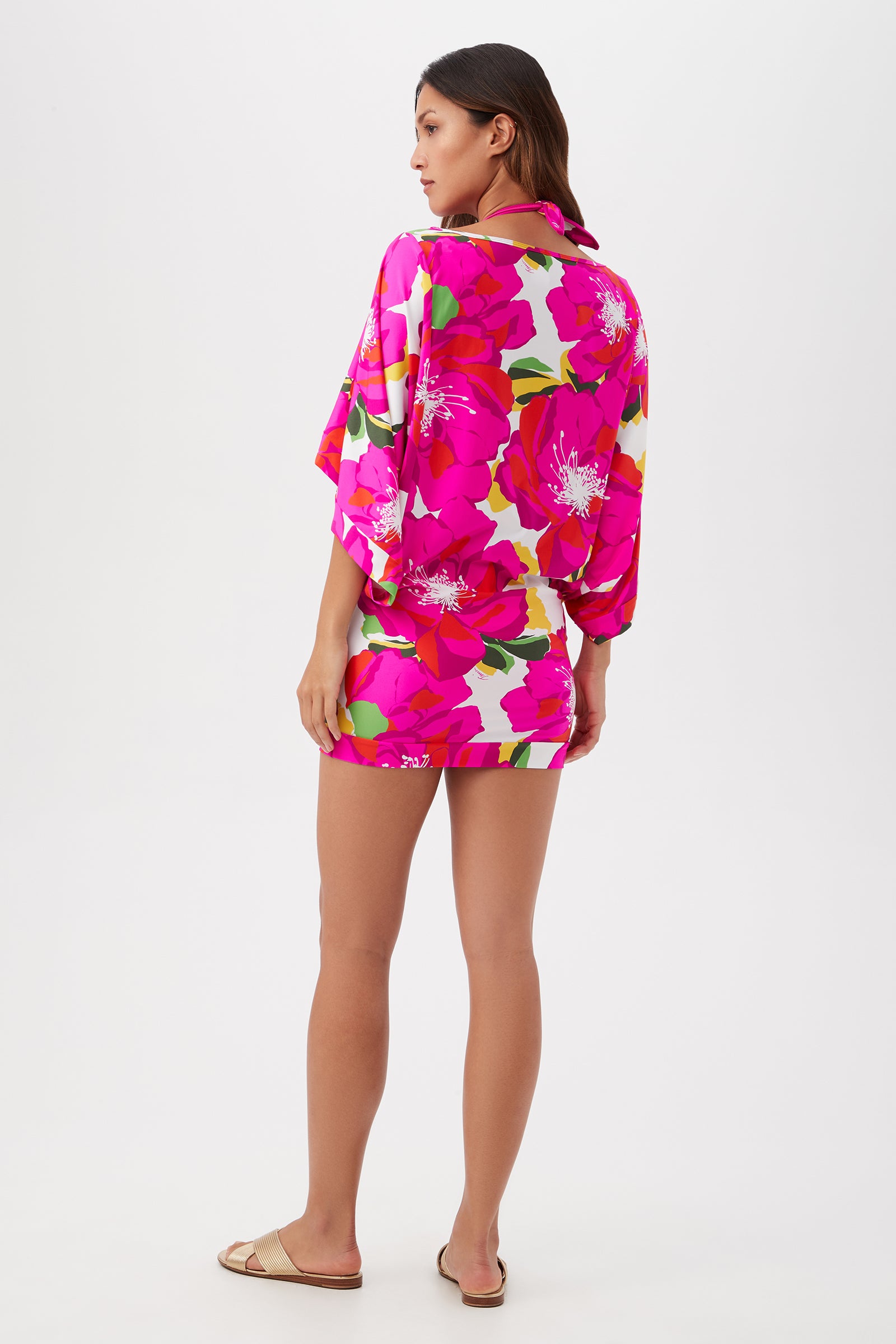 A woman faces away, barefoot, with straight hair, wearing the Bloom Casablanca Swim Tunic in bright pink floral print with a matching mini skirt against a plain white backdrop—a chic look perfect as a cover-up.
