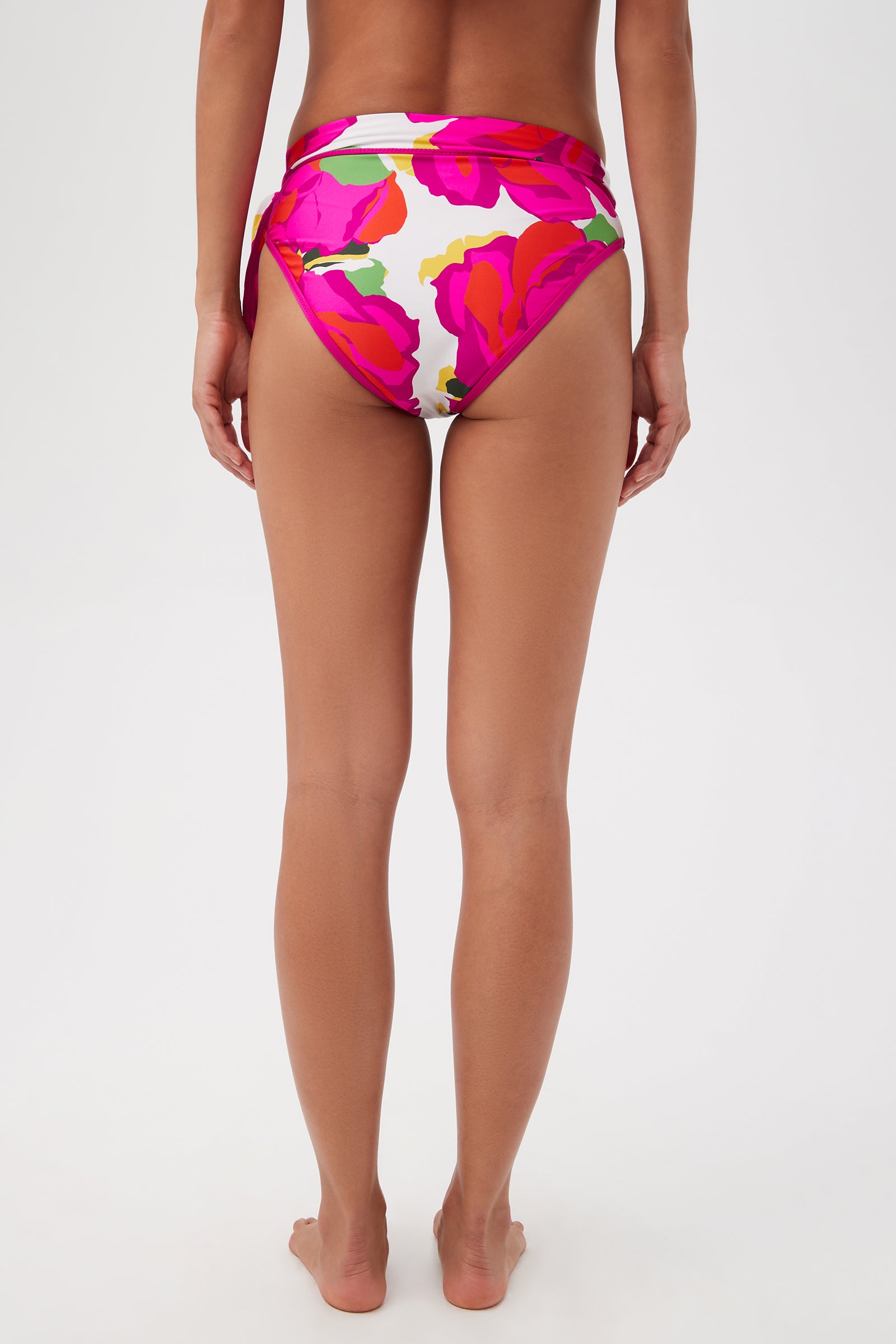 A person stands facing away, wearing the Bloom Sash High Waist bikini bottom in multi, featuring a bold pink and red floral pattern on white, full coverage, and an adjustable sash tie belt. The background is plain white.