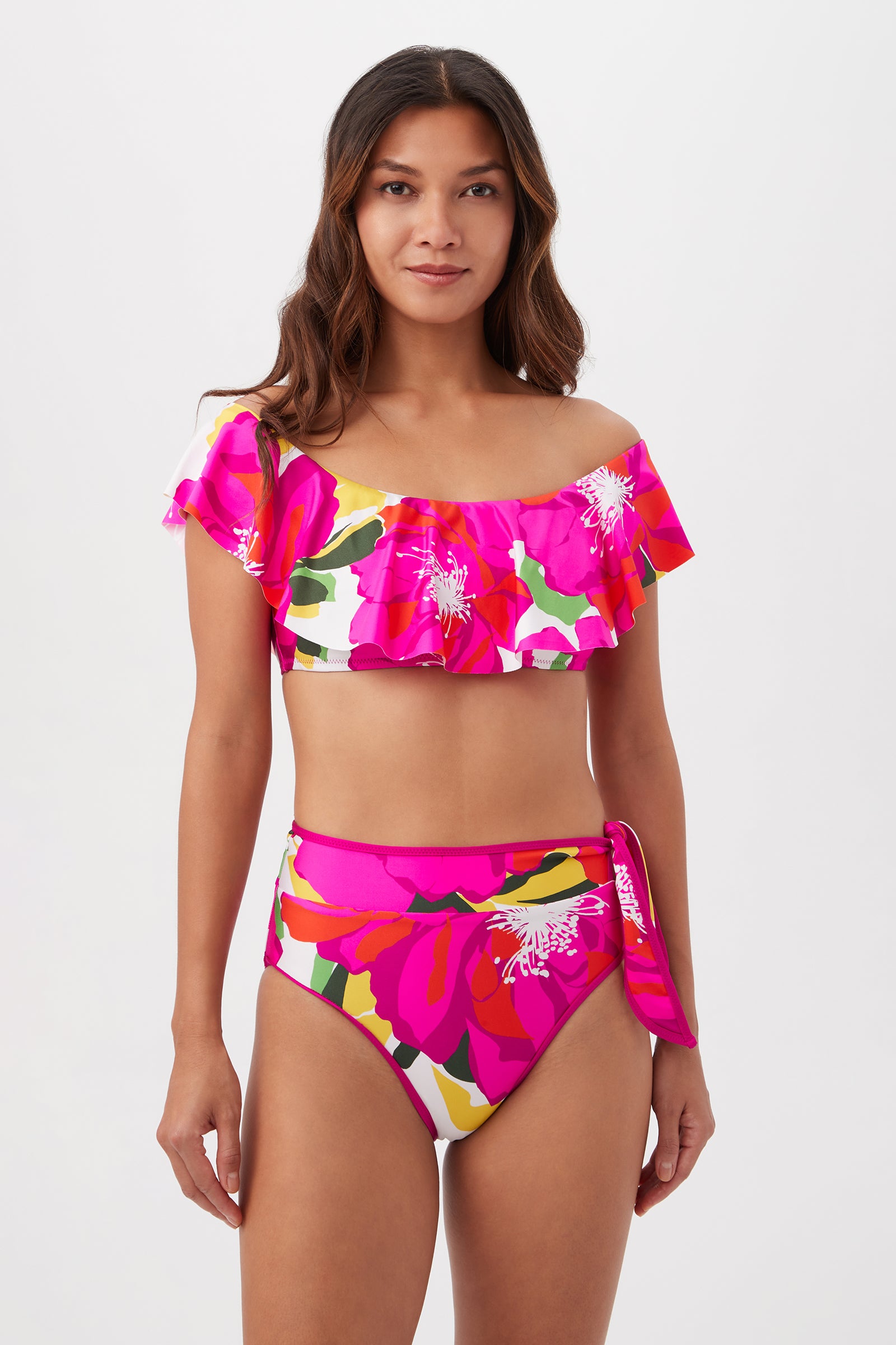 Model wears the Bloom Sash High Waist bikini, featuring a ruffled off-shoulder top and high-waist bottom with colorful pink, yellow, red, and white floral patterns. She poses against a light background with a slight smile.