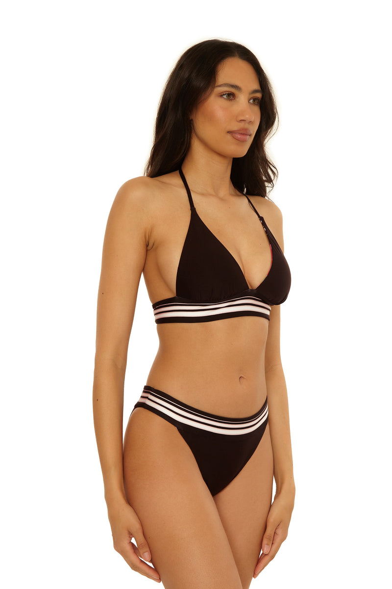 A woman models the Reversible Hipster Swim Bottom - Bougainvillea, featuring black fabric with white and pink striped bands, standing against a plain white background with a neutral expression, looking slightly to the side.