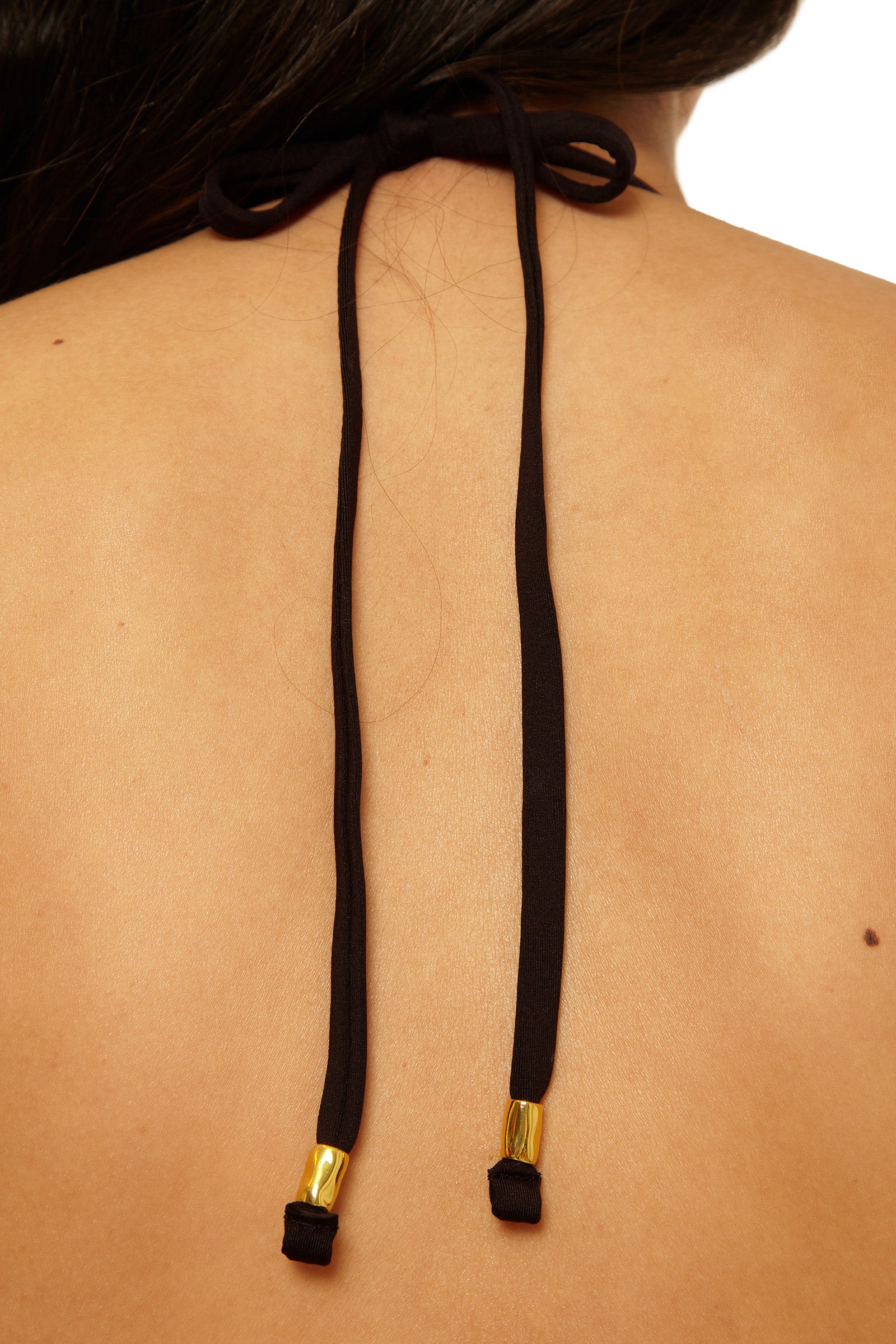 Close-up of a person’s upper back showing the tied black straps of the Tab Side Hipster Swim Bottom - Bougainvillea, featuring gold metal tips. The person’s hair partially rests on their back.