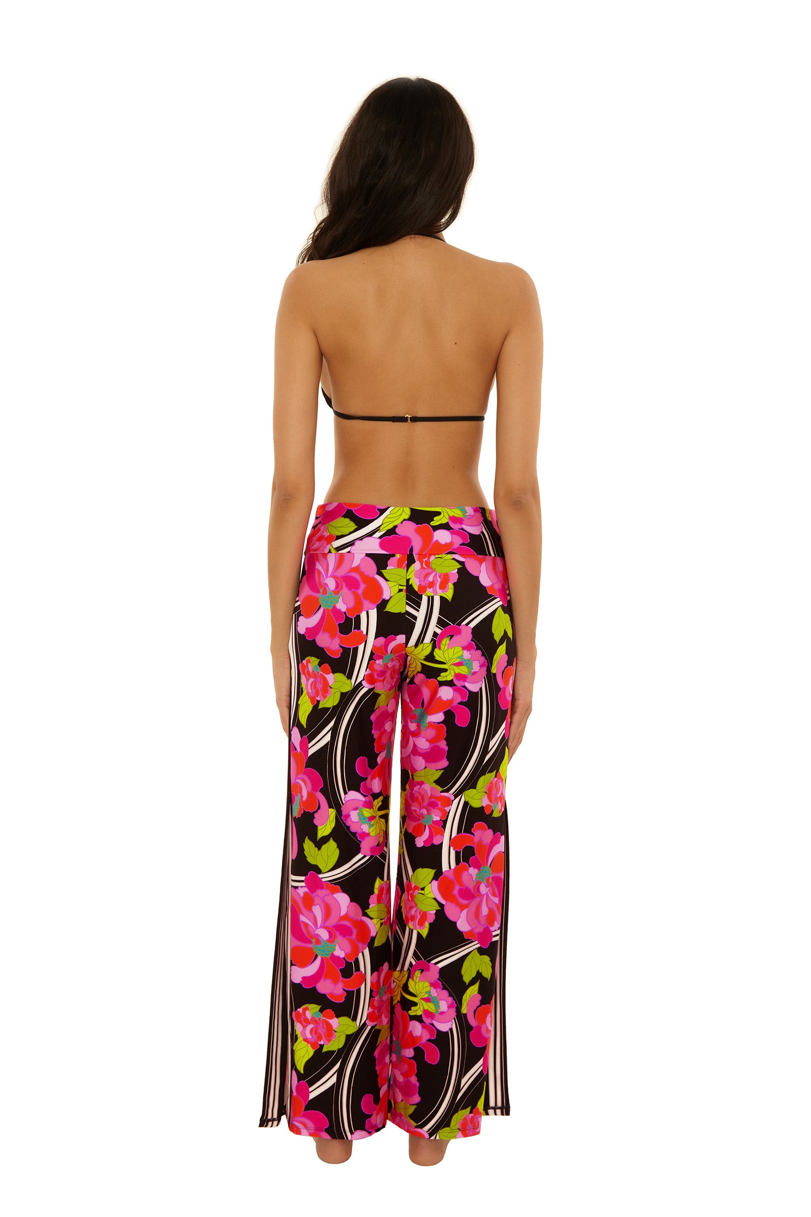 A woman stands barefoot with her back to the camera, wearing a black bikini top and the Swim Cover-Up Pant - Bougainvillea, featuring a vibrant pink, red, and green floral pattern on black.