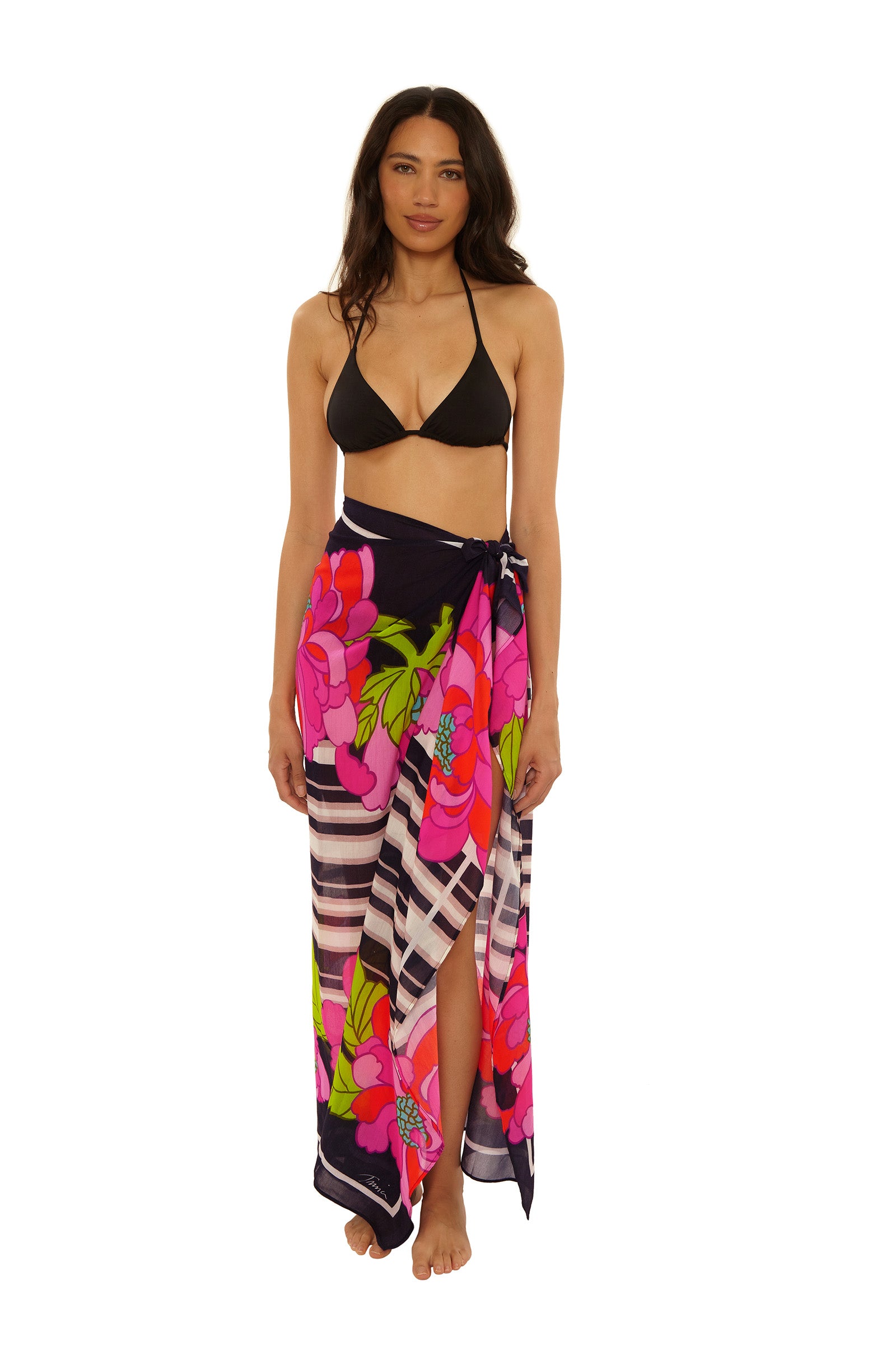 A woman, barefoot against a white background, wears a black bikini top with the Pareo Swim Cover-Up in Bougainvillea Swim Knit.