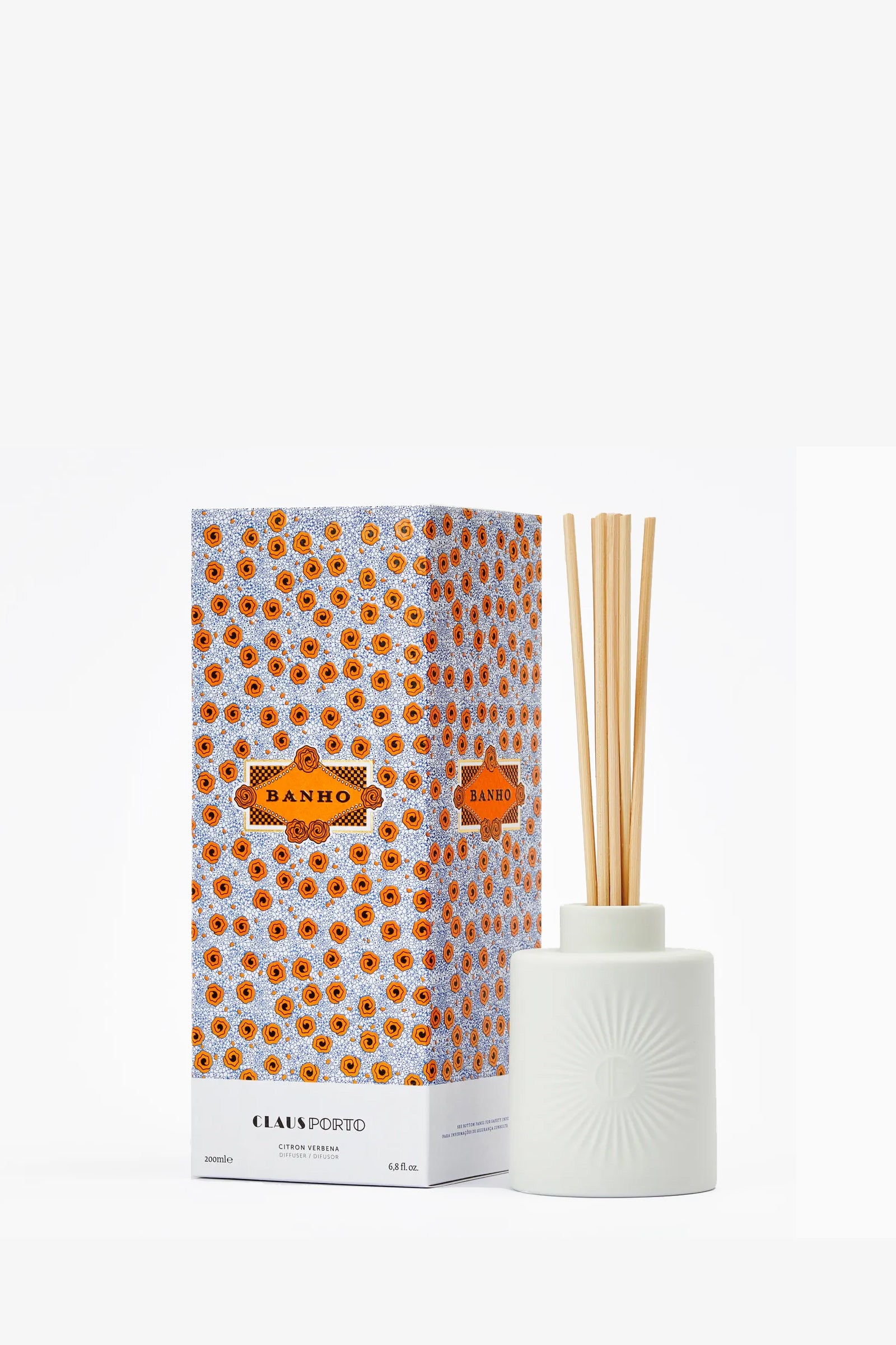 A white ceramic Claus Porto Banho Diffuser with reeds stands beside a decorative orange and blue patterned box labeled “Banho.”.