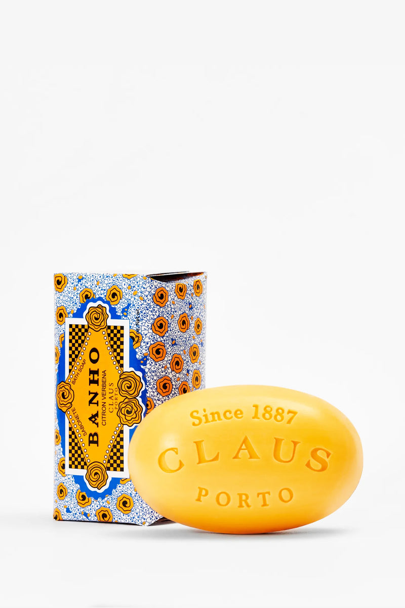 A yellow oval Claus Porto Banho Soap bar labeled “Since 1887” is displayed in front of its vibrant, floral-patterned box featuring the text “Banho” and “Citrus Verbena.”.