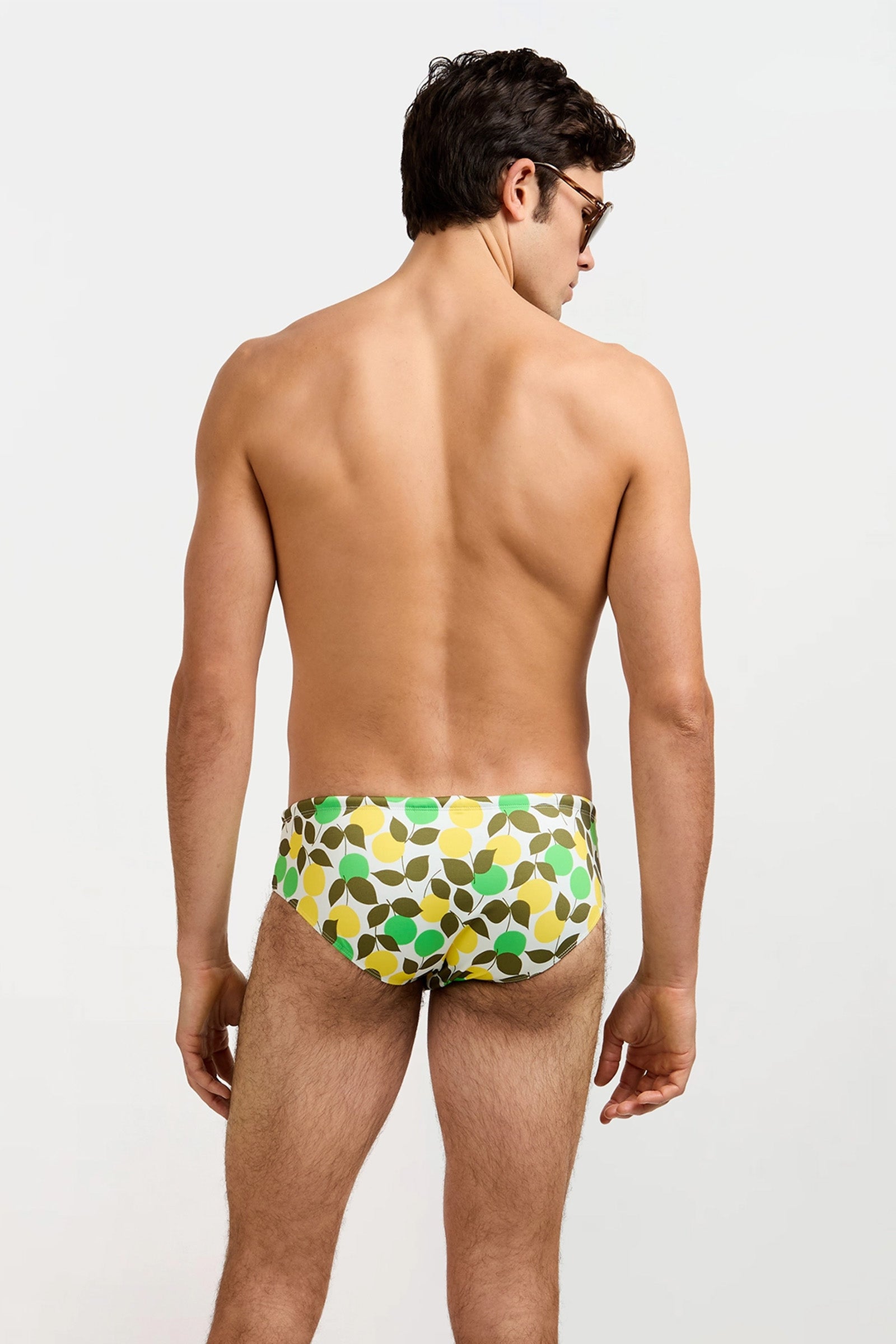 A man with short brown hair stands facing away, wearing sunglasses and the Bondi Swim Brief - Limone, which features green, yellow, and white leaf patterns on a light background.