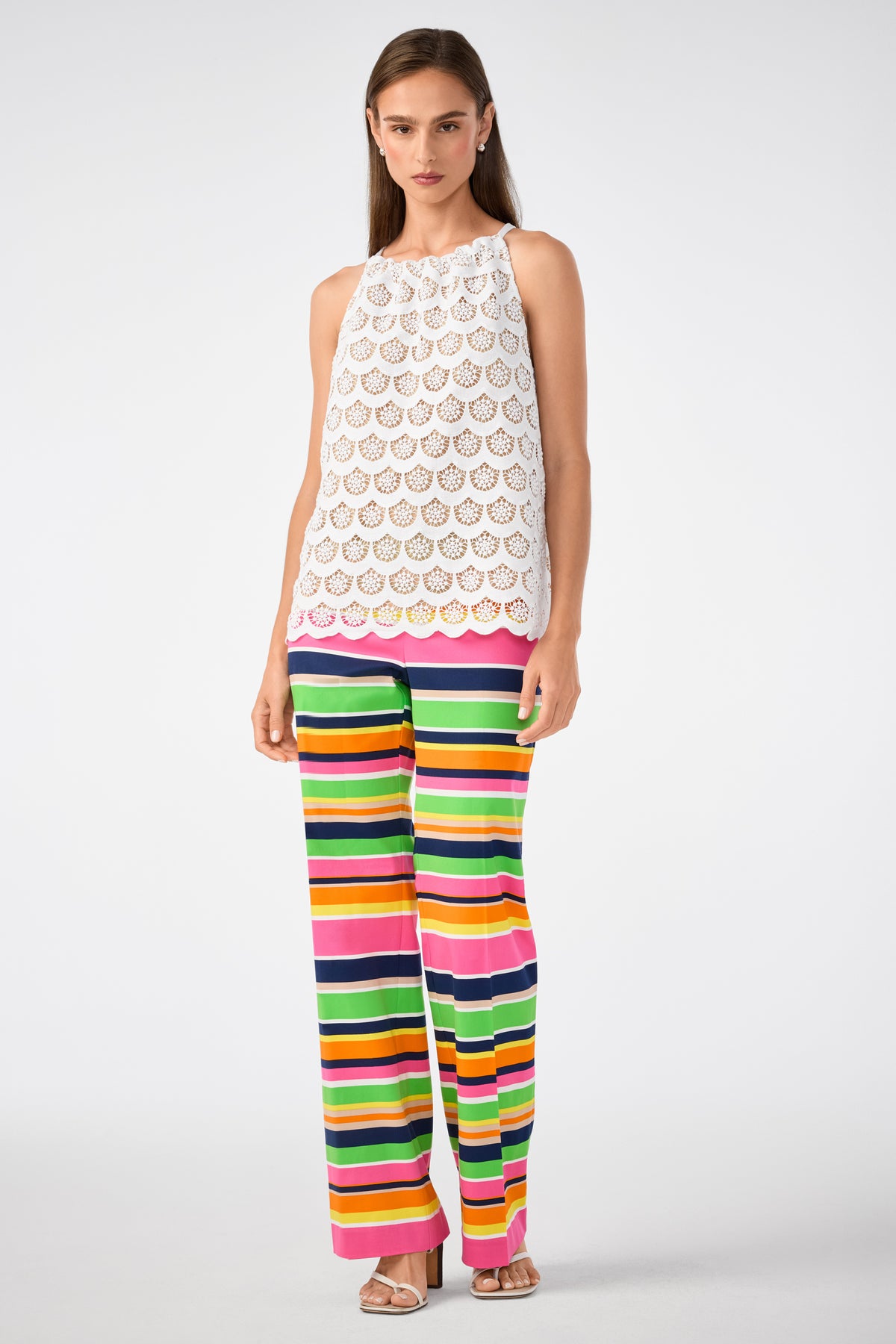 A woman with straight brown hair and sandals wears the Rancho Crochet Halter Top, a sleeveless white crochet halter, paired with high-waisted, wide-leg pants featuring bold horizontal stripes in green, yellow, pink, black, and orange.