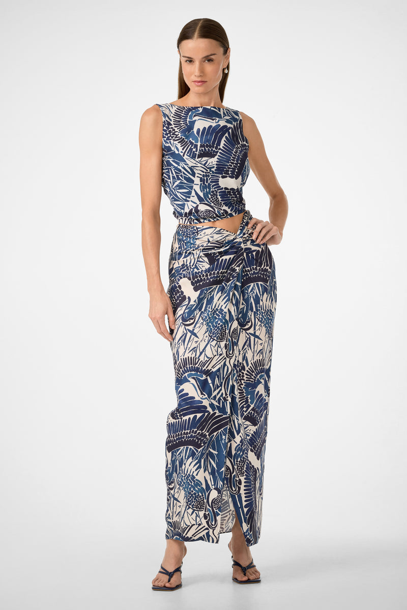 A woman models the Alodia Tie-Front Maxi Skirt - Tsuru, a blue and white printed, high-waisted, ankle-length skirt with a side slit. She pairs it with a matching sleeveless top and black sandals against a plain white background.