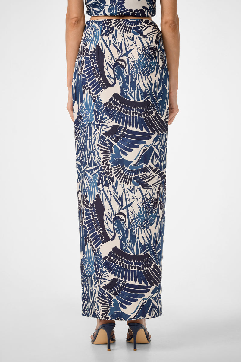 A woman is seen from the back wearing the Alodia Tie-Front Maxi Skirt - Tsuru, featuring a blue and white bird and foliage print, paired with dark heeled sandals against a plain white background.