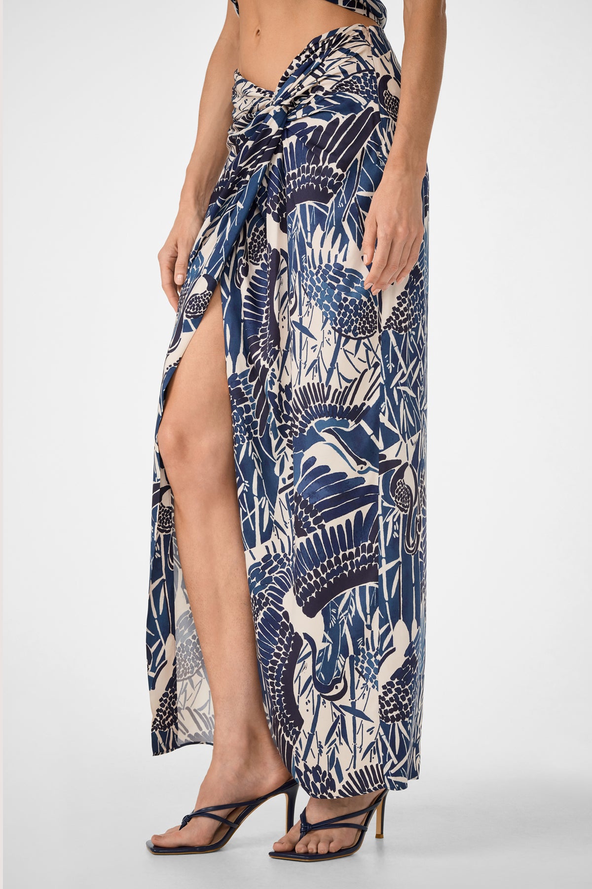 Wearing the Alodia Tie-Front Maxi Skirt - Tsuru, a navy blue and cream sarong-style skirt with an abstract bird and foliage print, high slit, and waist tie, paired with black high-heeled sandals.