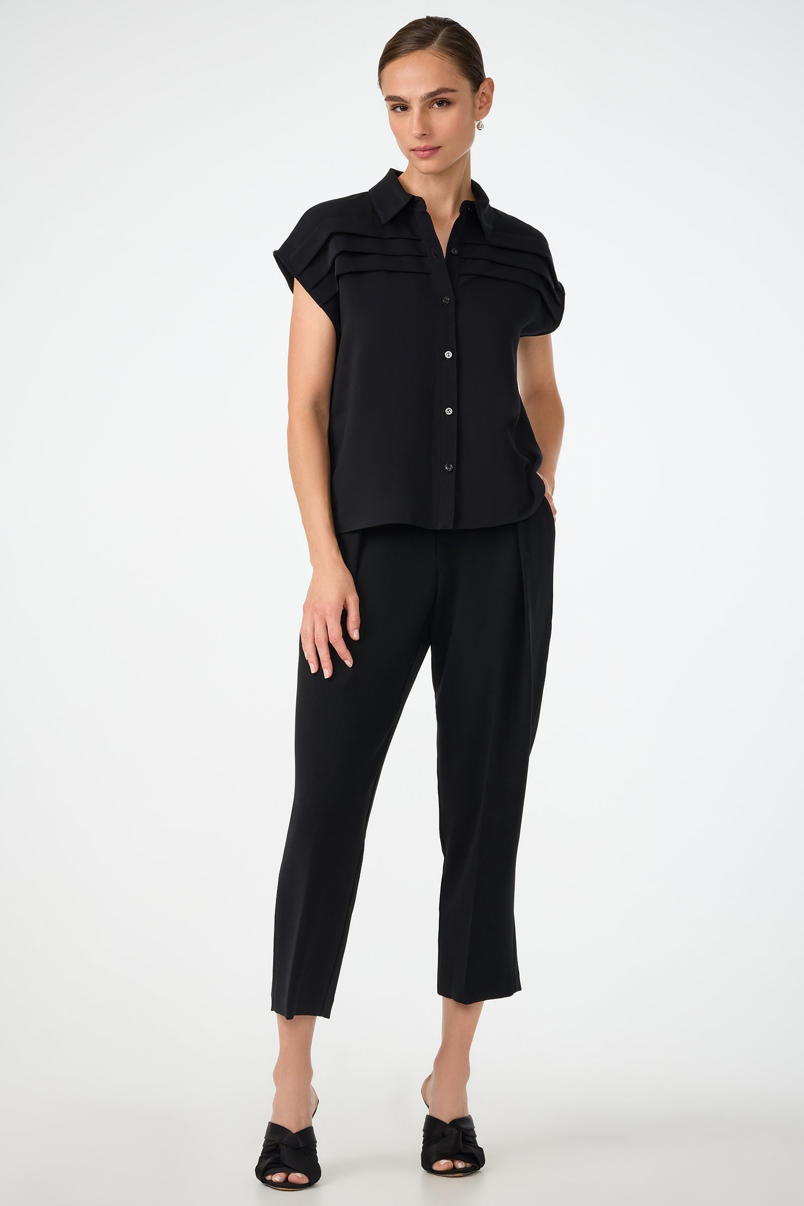 A woman stands against a plain background wearing the Cheri Pleated Button-Front Top, a short-sleeve black blouse with pleat details, paired with cropped black pants and black heeled sandals. Her hair is pulled back.