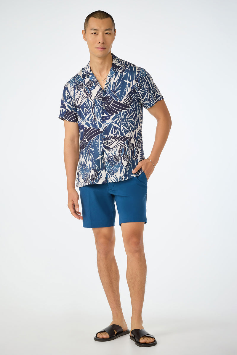 A man stands against a plain background wearing the Travis Short in Sumi Blue, paired with a blue-and-white patterned shirt, black slide sandals, and a bracelet, with one hand in his pocket.